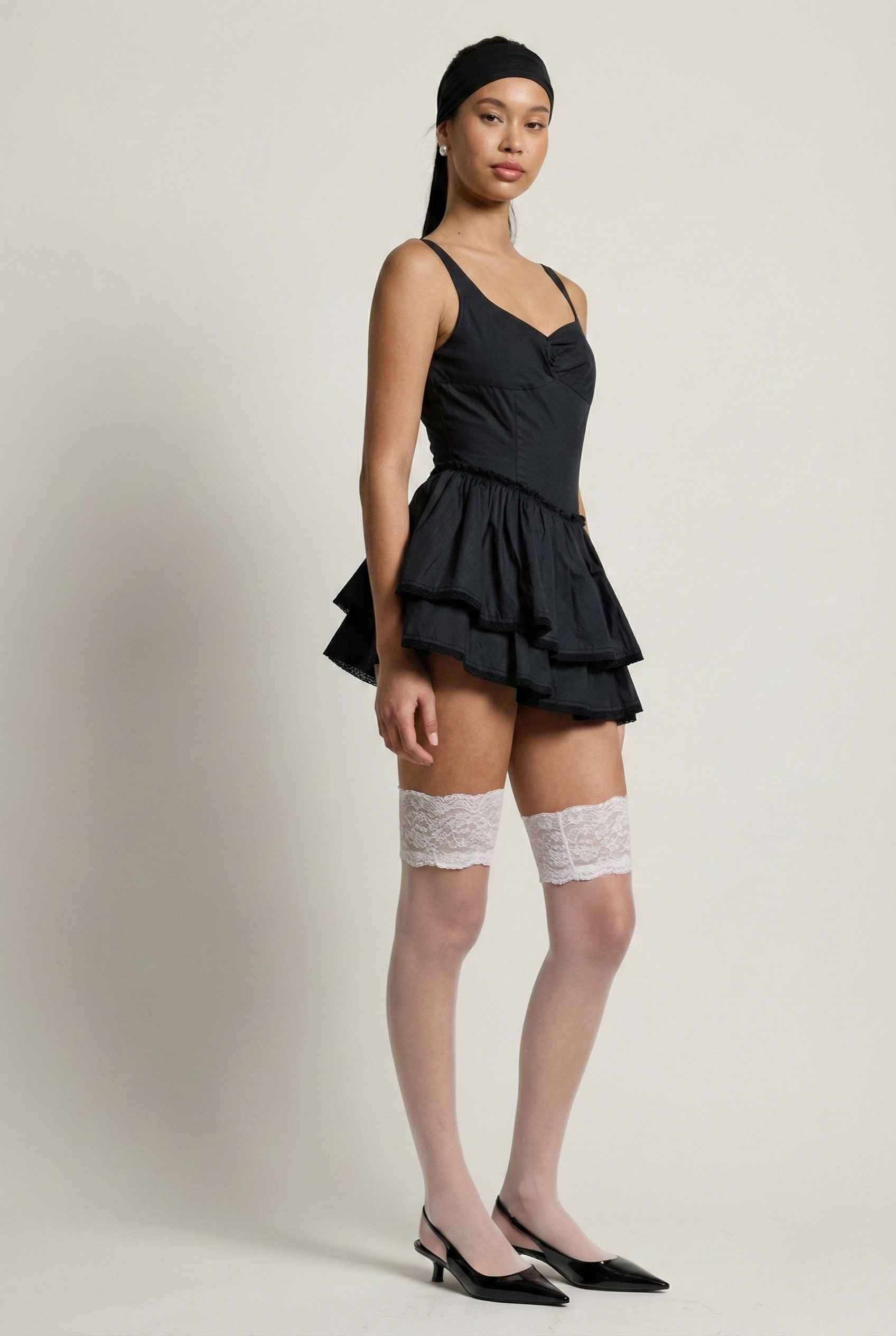 Woman wearing a Elodie Mini Black Tiered Ruffle Cotton Mini Dress with ruffled layers and knee-high socks on a plain background
