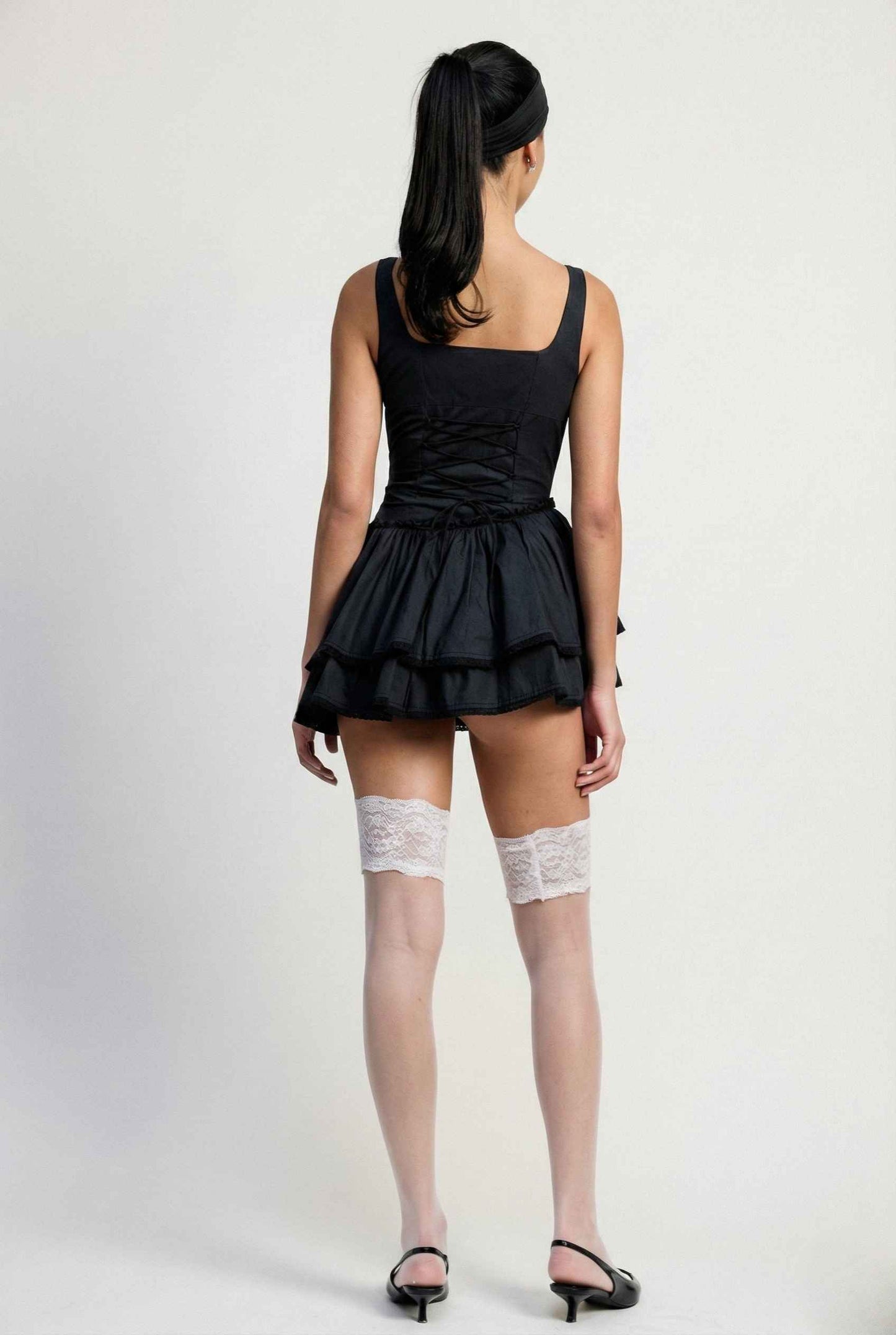 Woman wearing a Elodie Mini Black Tiered Ruffle Cotton Mini Dress with stockings on a plain background