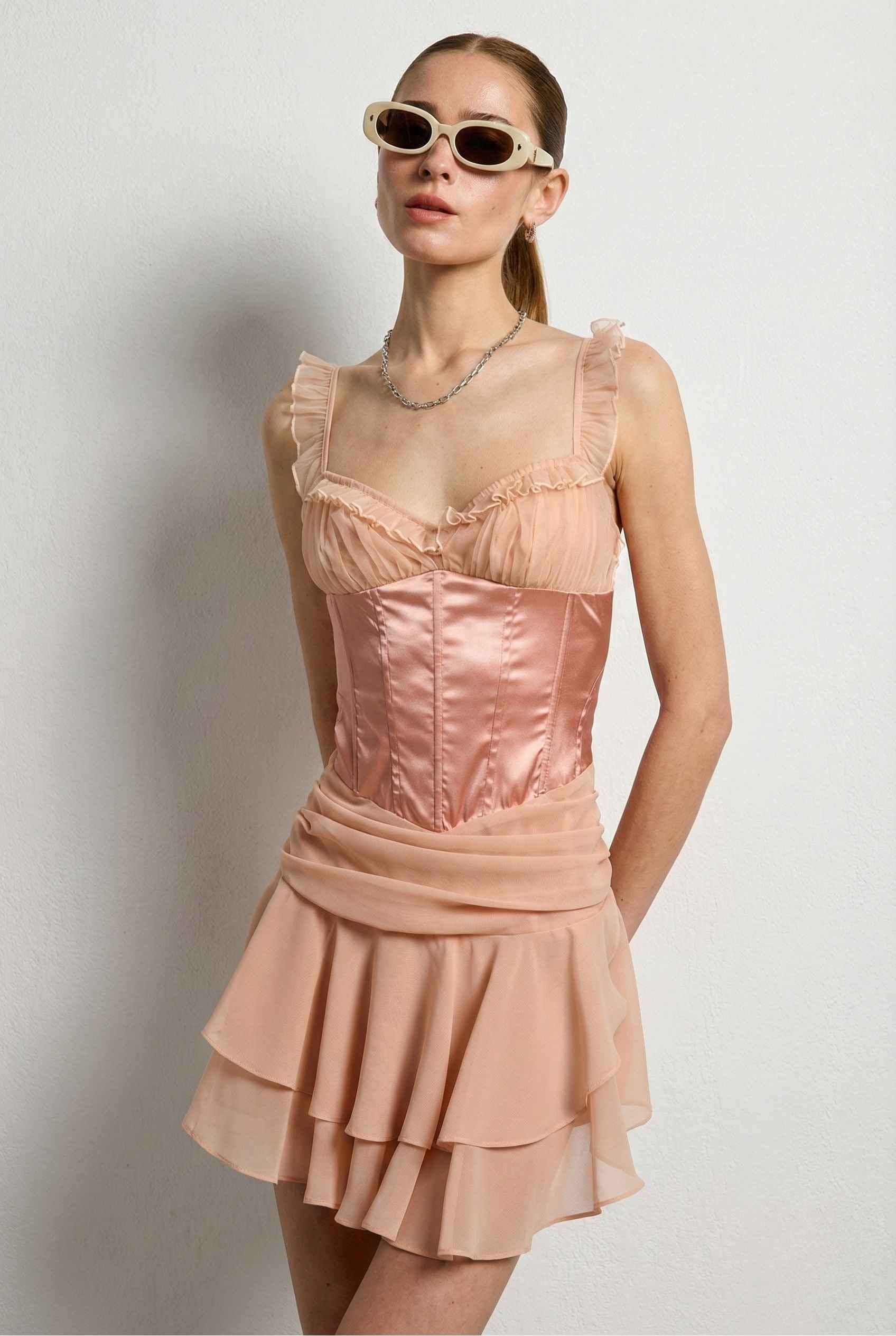 Woman wearing a Elodie Mini Pink Satin & Mesh Ruffle Bustier Dress with sheer overlay against a plain background