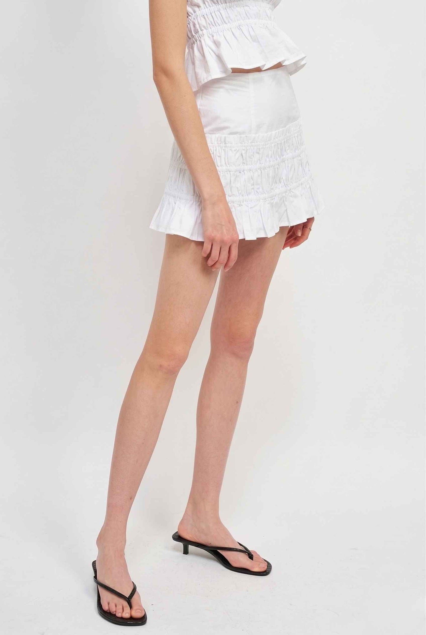 Woman wearing a Elodie Skirt White Cotton Smocked Ruffle Mini Skirt on a plain background