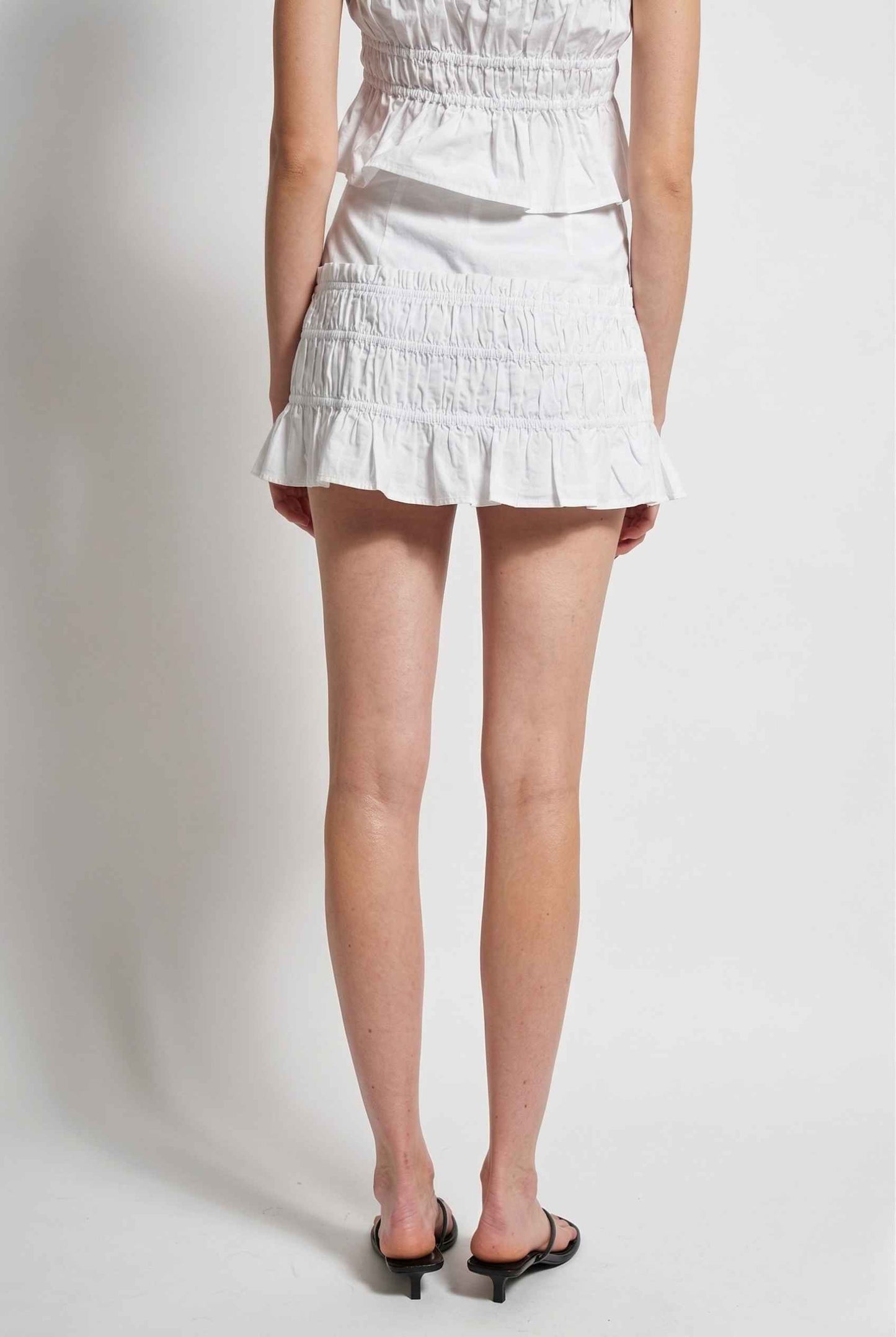 Elodie Skirt White Cotton Smocked Ruffle Mini Skirt worn by a woman on a plain background