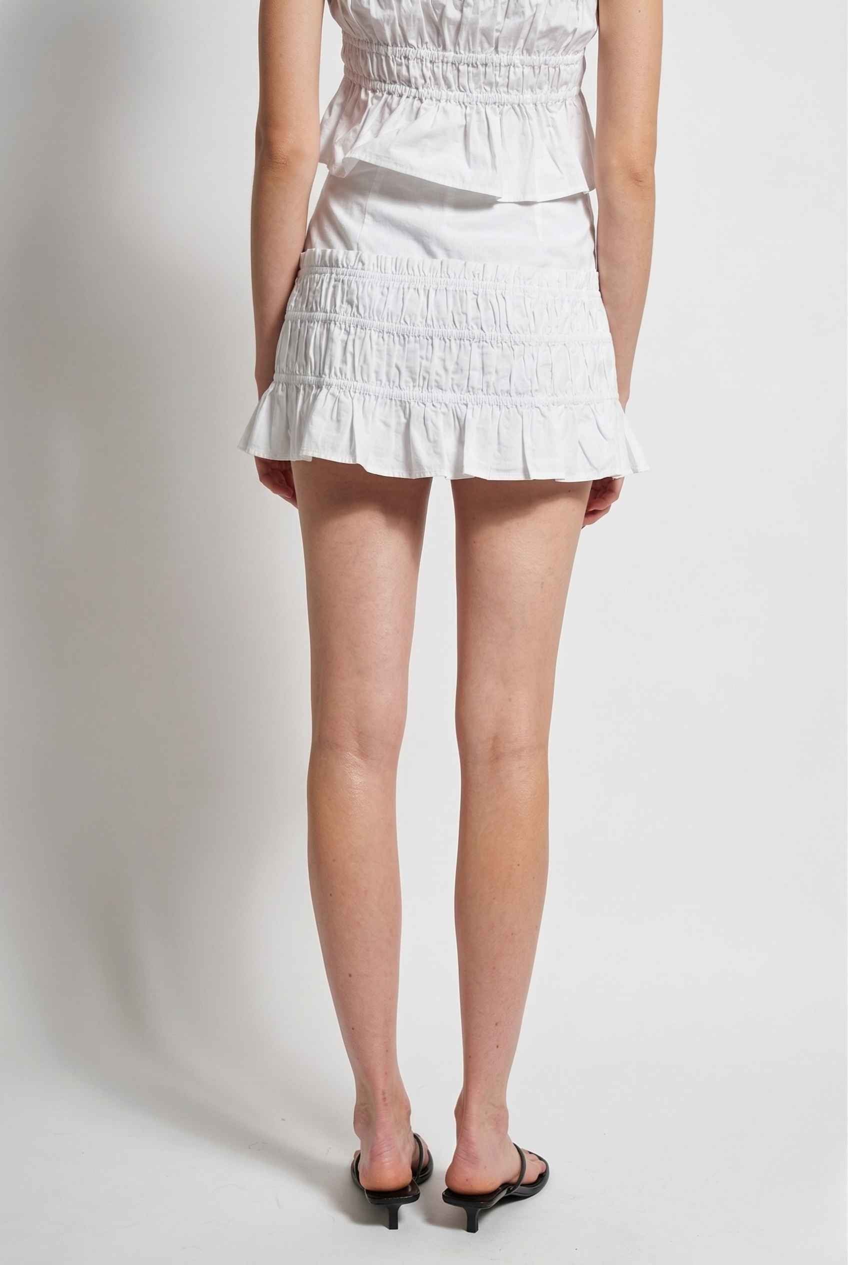 Elodie Skirt White Cotton Smocked Ruffle Mini Skirt worn by a woman on a plain background