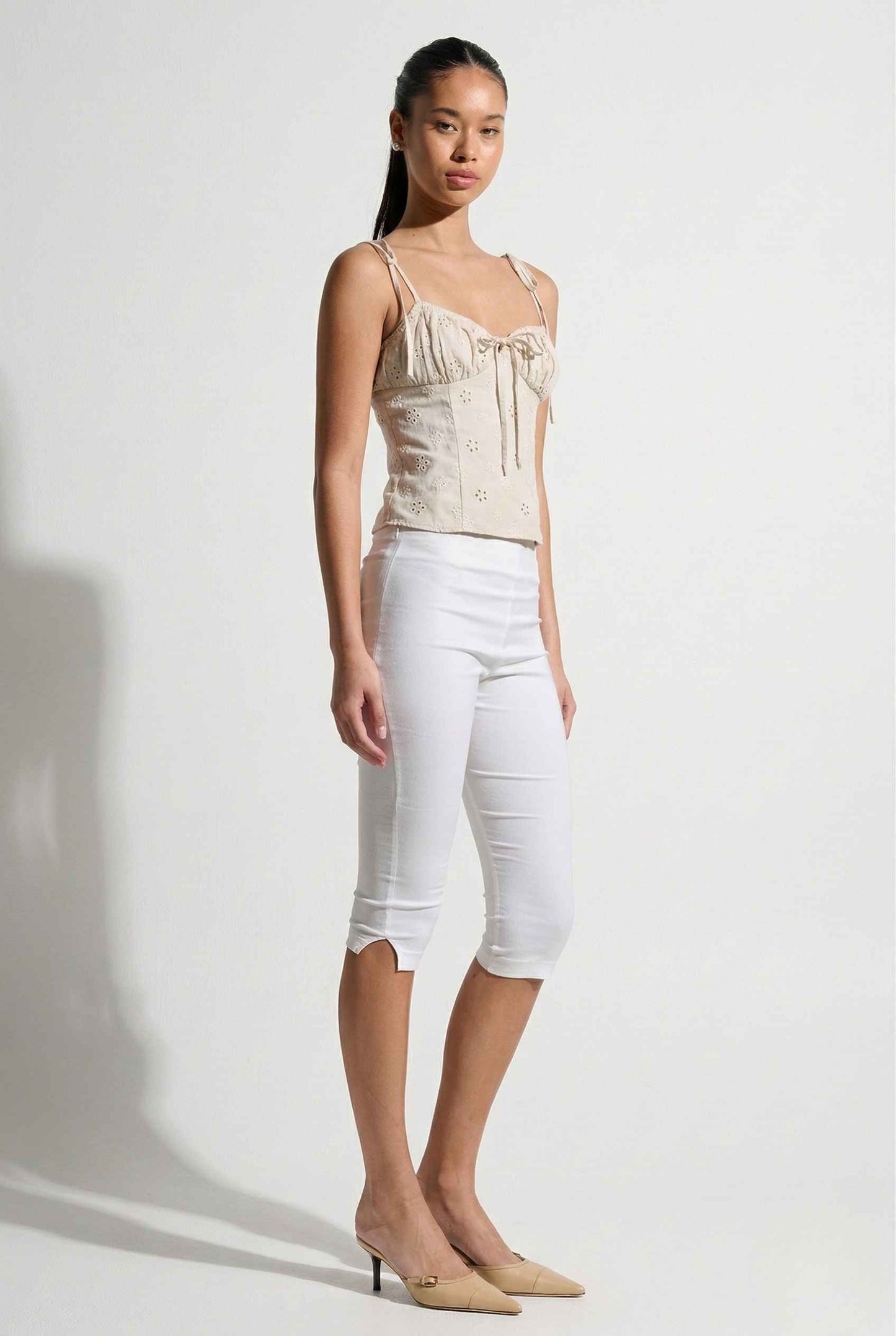 Woman wearing a Elodie Top Ivory Floral Embroidered Tie-Strap Corset Top and white capri pants on a plain background