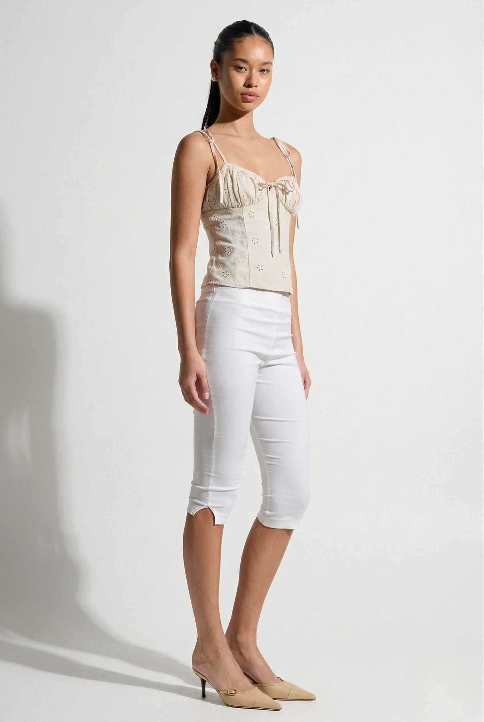 Woman wearing a Elodie Top Ivory Floral Embroidered Tie-Strap Corset Top and white capri pants on a plain background