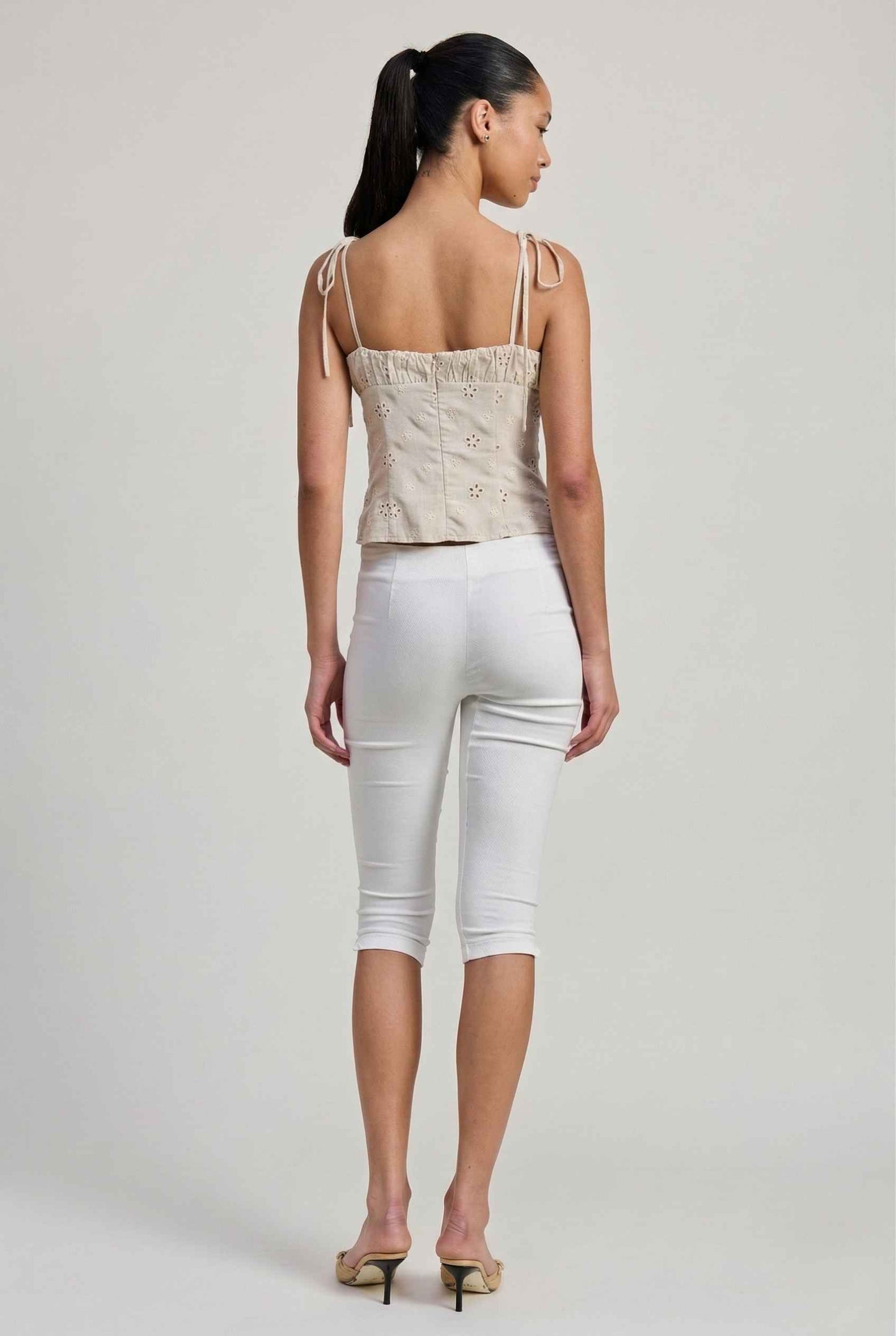 Woman wearing a Elodie Top Ivory Floral Embroidered Tie-Strap Corset Top and white capri pants on a plain background