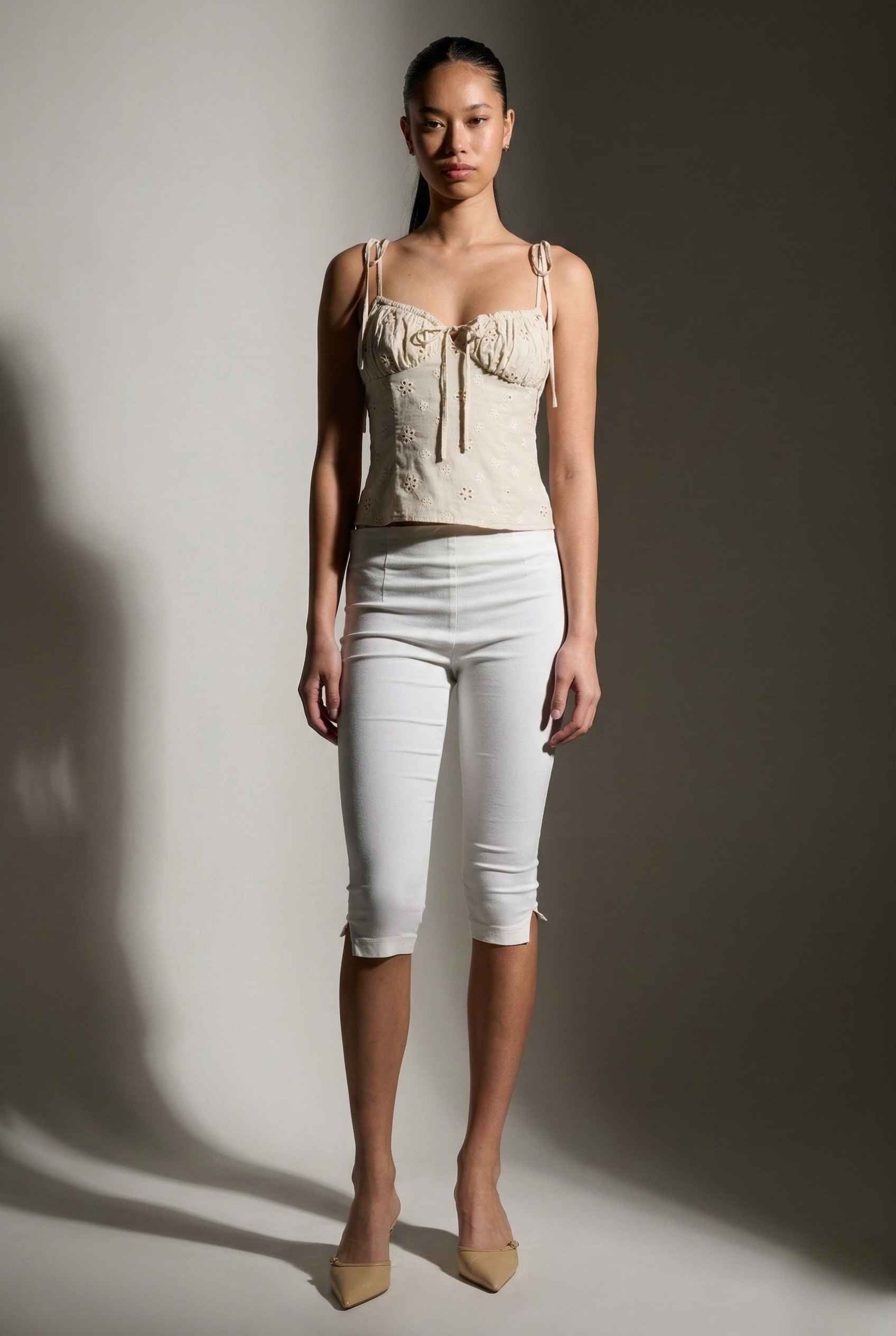 Woman wearing a Elodie Top Ivory Floral Embroidered Tie-Strap Corset Top and white pants against a plain background