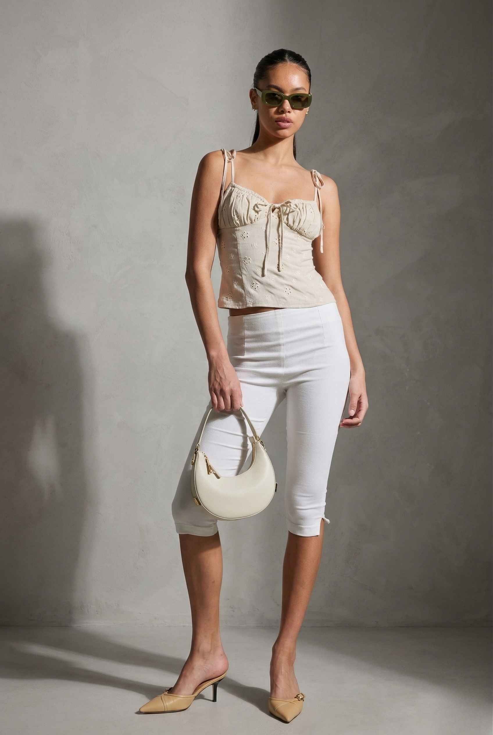 Woman wearing a Elodie Top Ivory Floral Embroidered Tie-Strap Corset Top and white pants, holding a cream handbag against a gray background