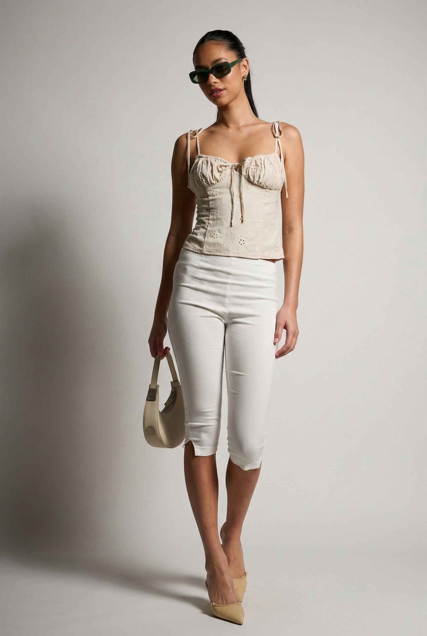 Woman wearing a Elodie Top Ivory Floral Embroidered Tie-Strap Corset Top and white pants with sunglasses and a handbag on a plain background