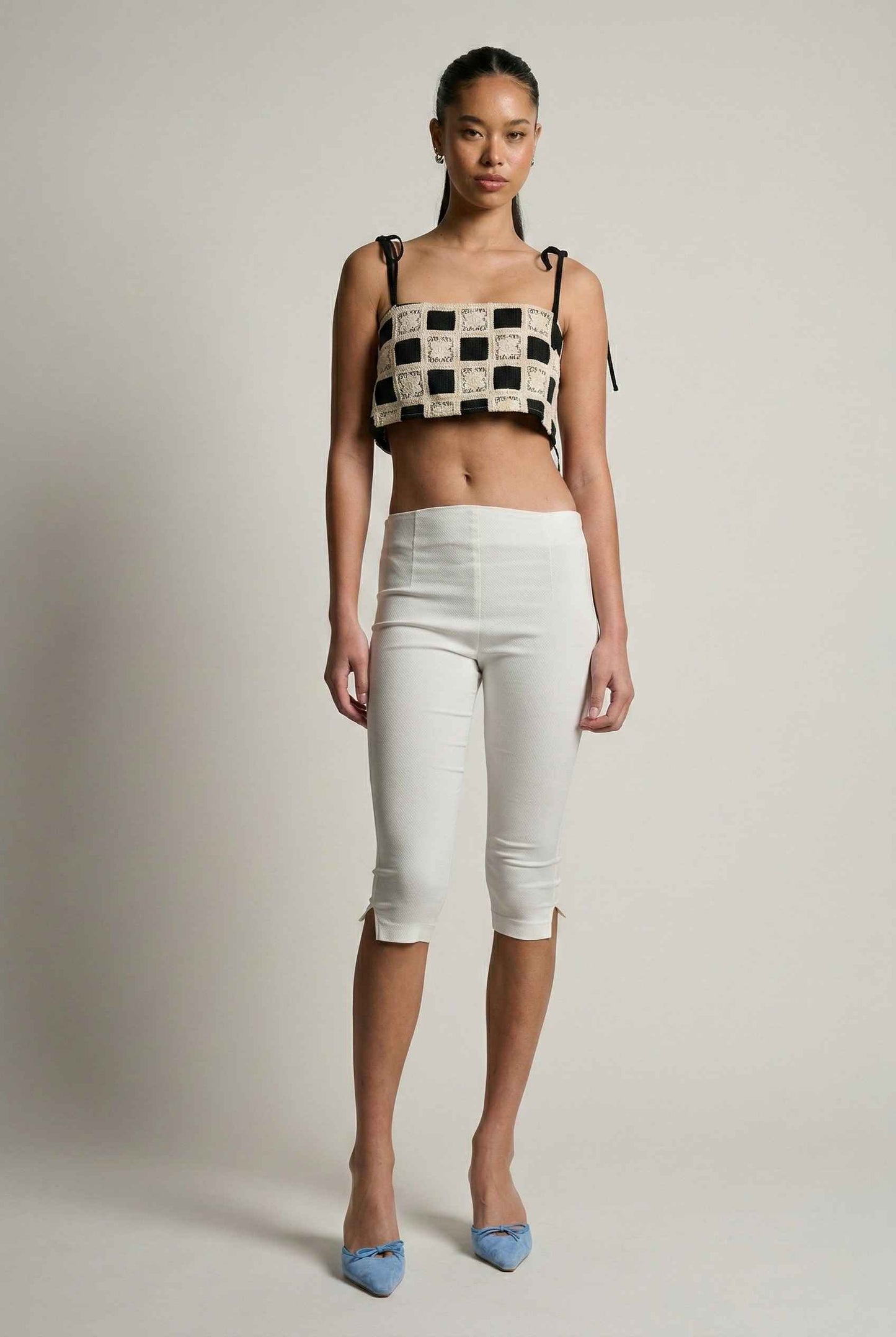 Model wearing Elodie Top in Black Jacquard Square Neck Cami with Ribbon Tie Straps with capri pants and heels on a plain background