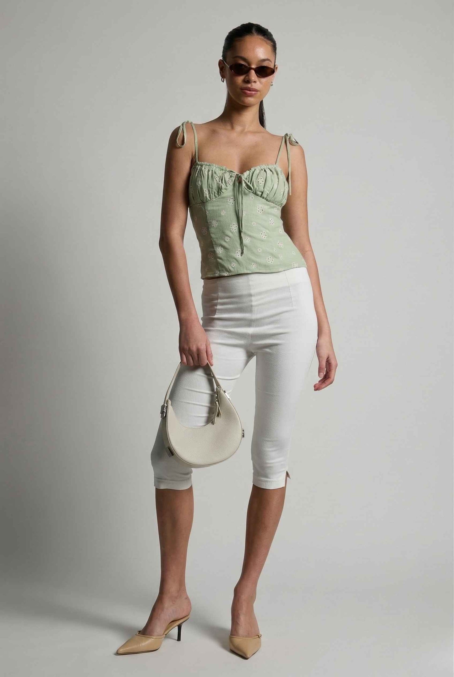 Woman wearing a Elodie Top Mint Floral Embroidered Tie-Strap Corset Top and white pants holding a cream handbag against a plain background