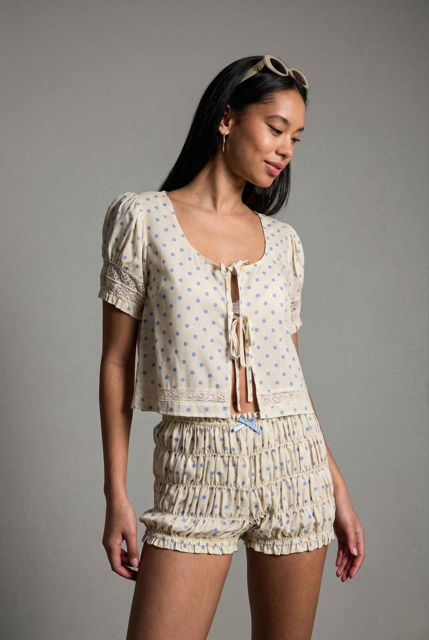 Woman wearing a Elsie Top Ivory & Blue Polka Dot Linen Tie-Front Blouse and shorts set on a gray background