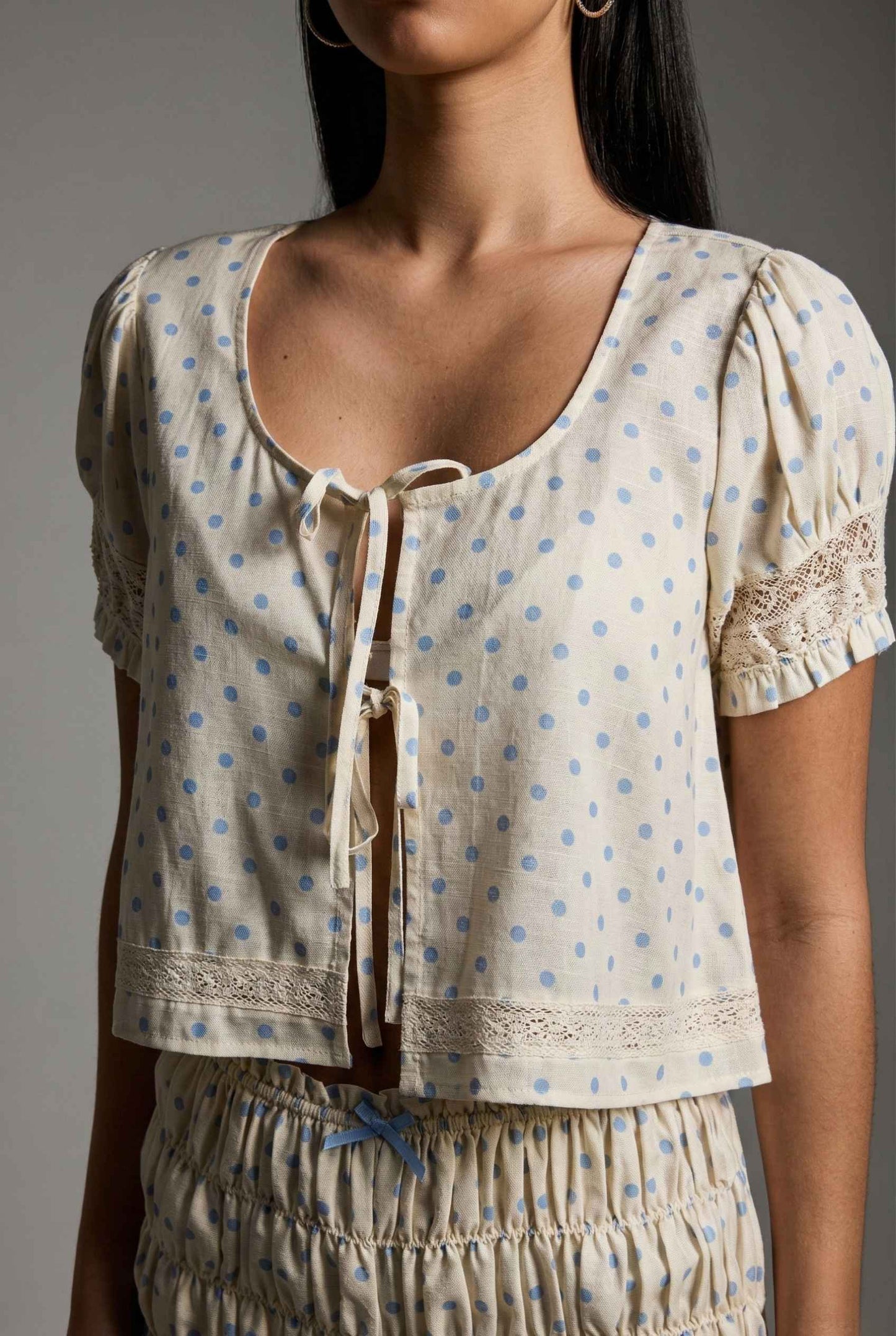 Woman wearing a Elsie Top Ivory & Blue Polka Dot Linen Tie-Front Blouse with lace details on a plain background