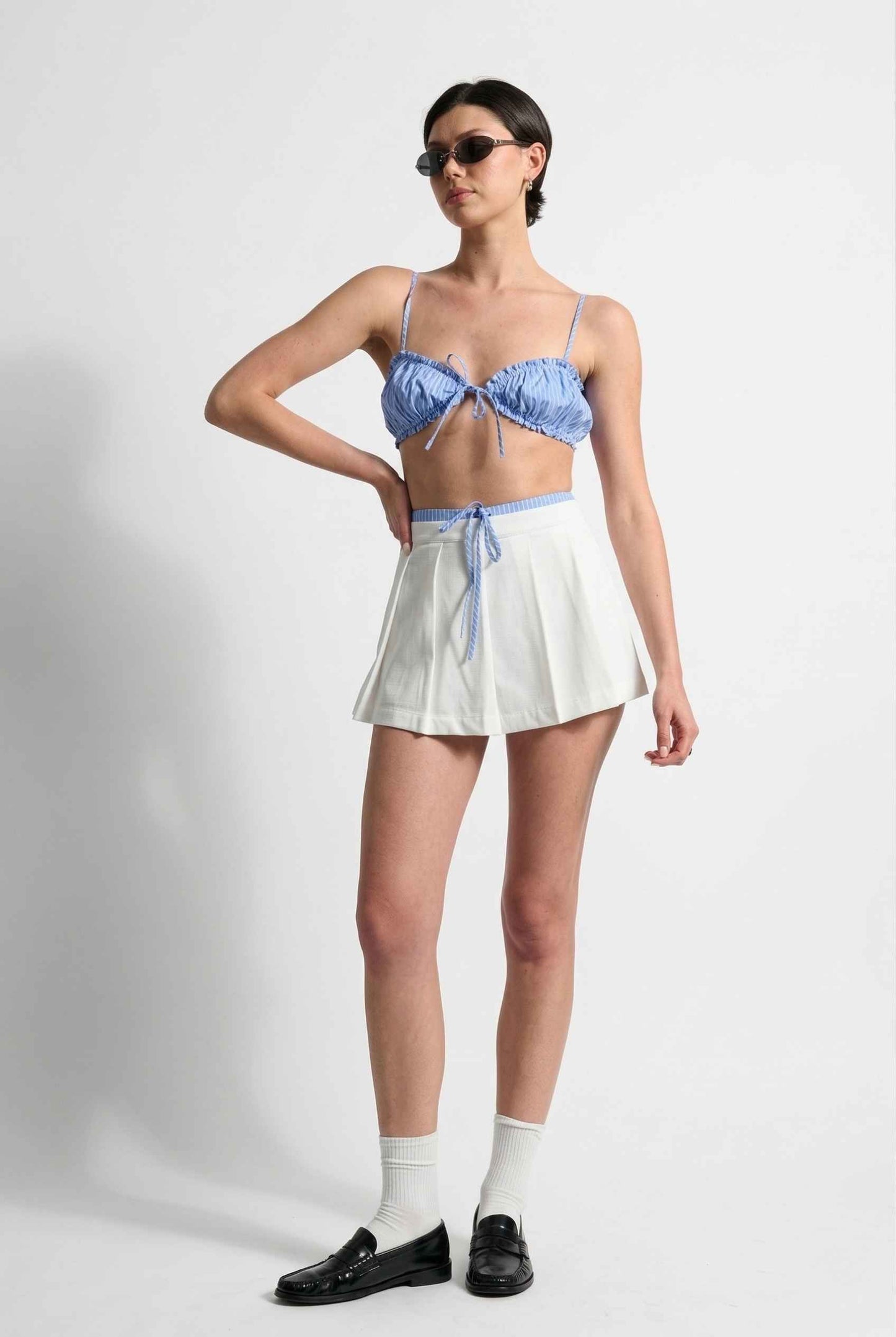 Model wearing Emory Skort in White Pleated Mini Skort with Striped Waist paired with matching bra top and shiny loafers with tall socks