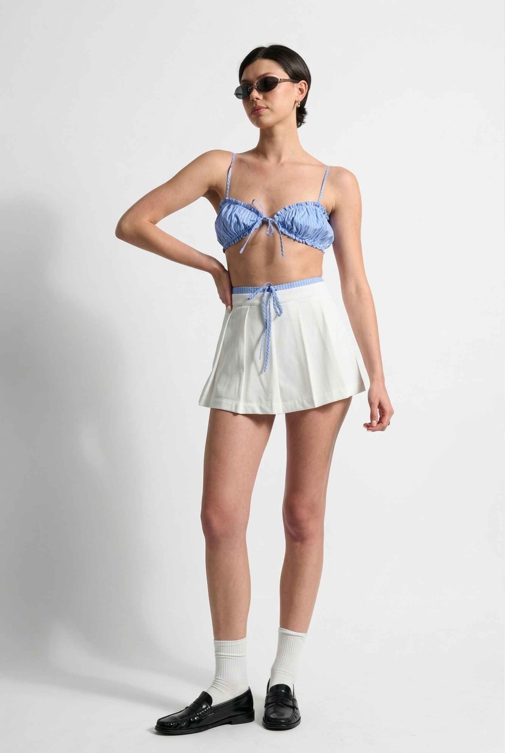 Model wearing Emory Skort in White Pleated Mini Skort with Striped Waist paired with matching bra top and shiny loafers with tall socks