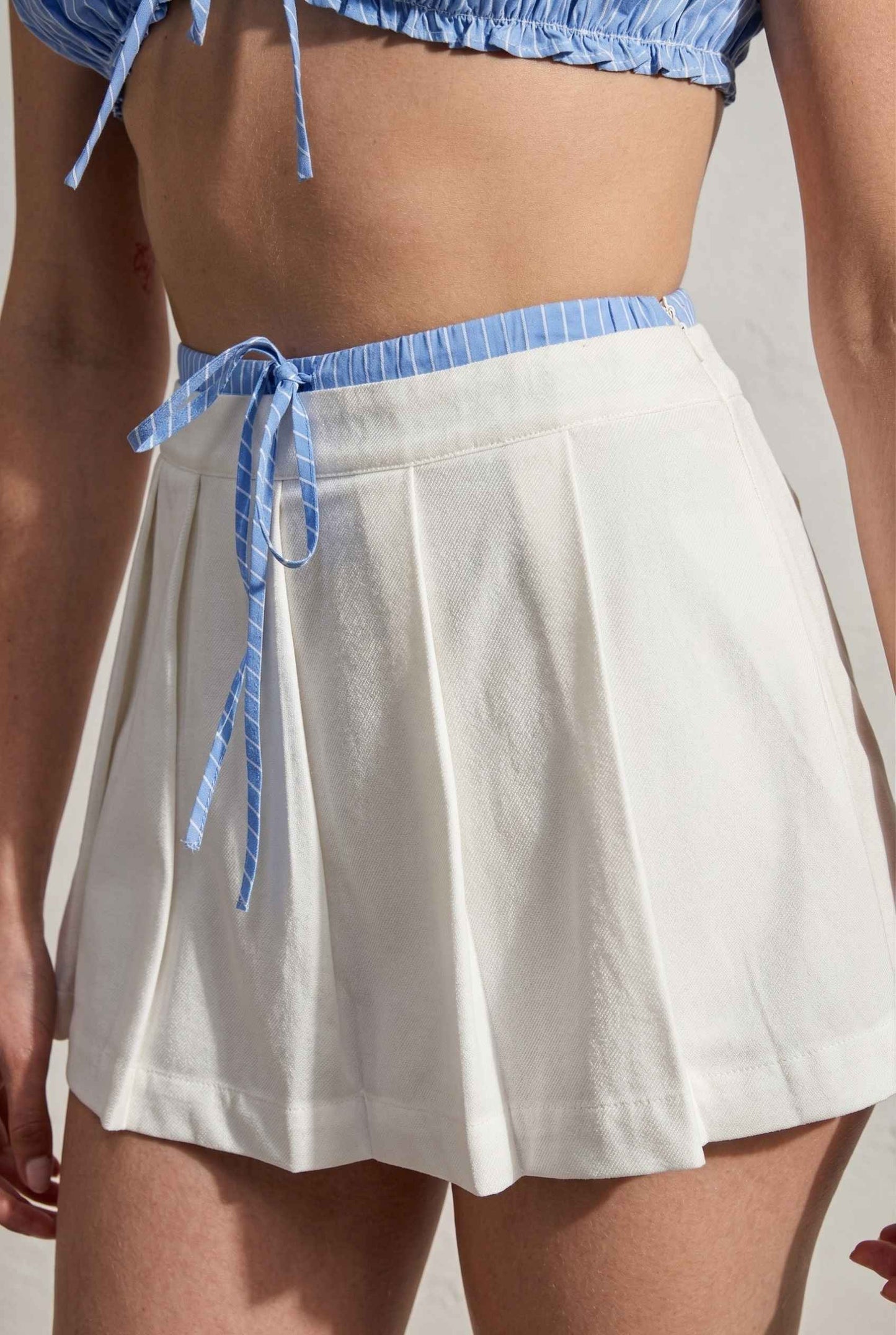 Model wearing Emory Skort in White Pleated Mini Skort with Striped Waist paired with matching bra top close up tied striped detail