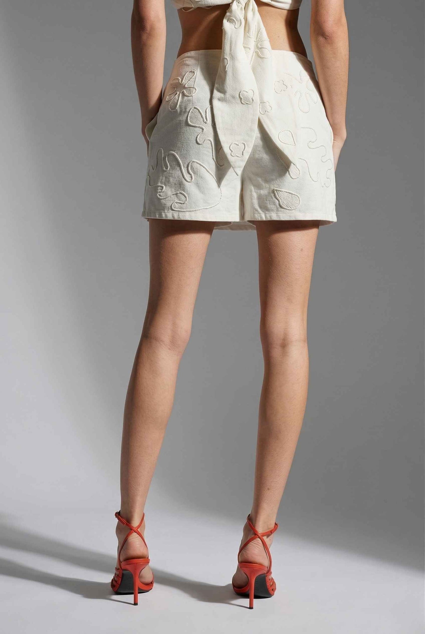 Woman wearing a Emory Skort White Tonal Embroidered Cotton-Linen Mini Skort with red high-heeled shoes on a gray background