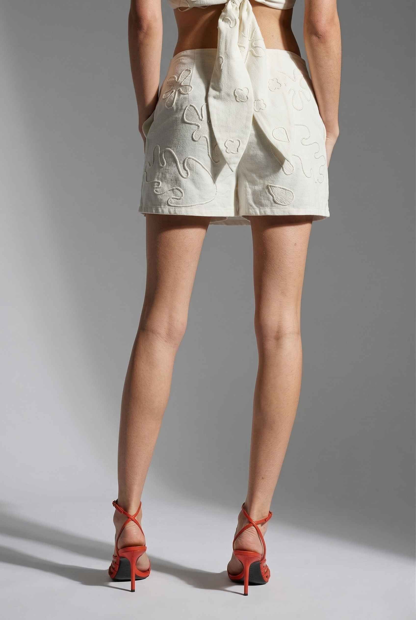 Woman wearing a Emory Skort White Tonal Embroidered Cotton-Linen Mini Skort with red high-heeled shoes on a gray background