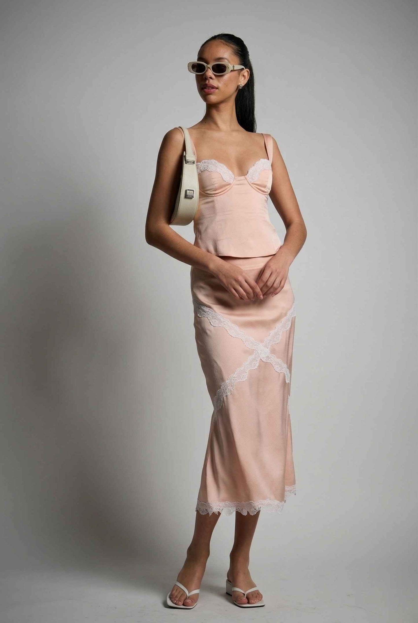 Woman wearing a Freya Skirt Blush Pink Satin Midi Skirt with White Lace Detail and sunglasses against a plain background