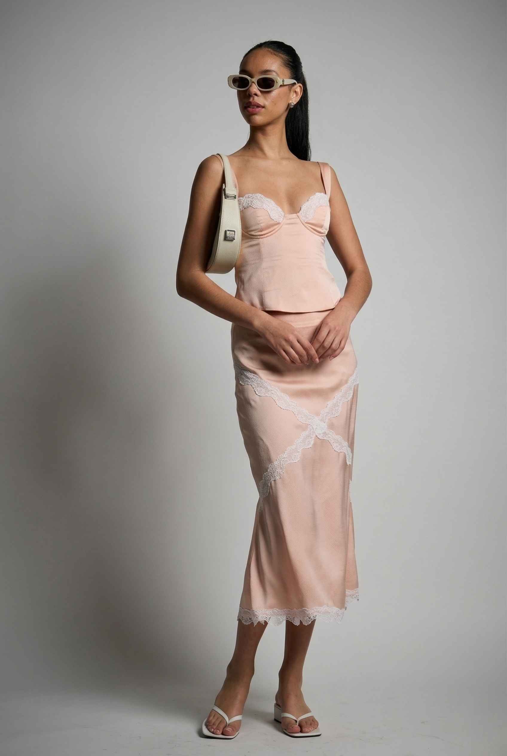 Woman wearing a Freya Skirt Blush Pink Satin Midi Skirt with White Lace Detail and sunglasses against a plain background