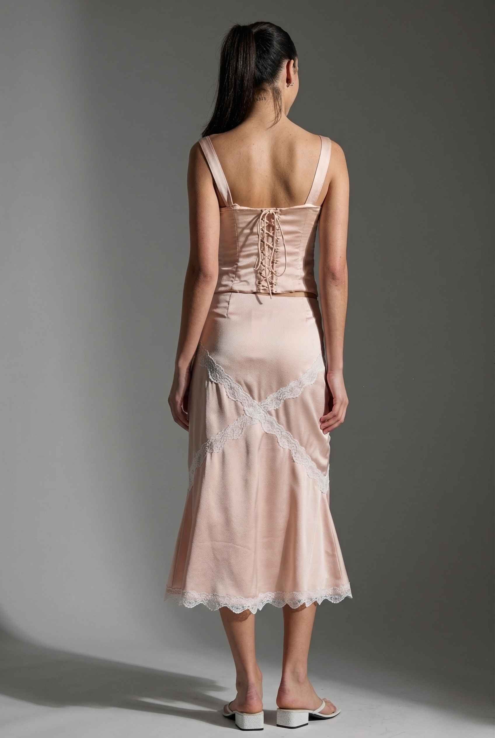 Woman wearing a Freya Skirt Blush Pink Satin Midi Skirt with White Lace Detail with lace details on a gray background