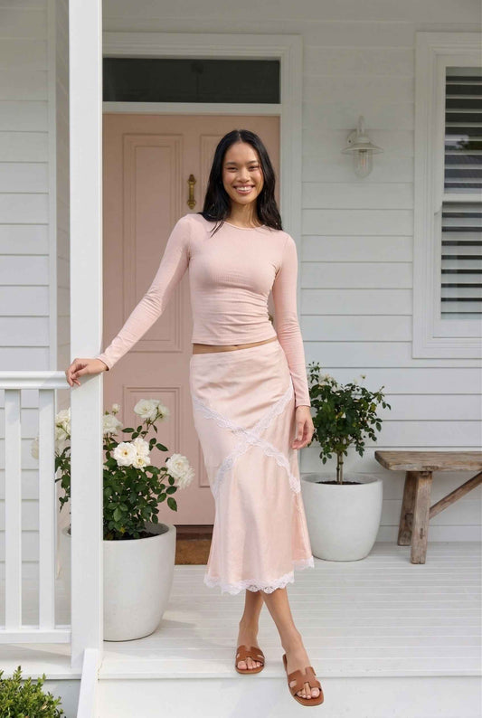 Woman in a Freya Skirt Blush Pink Satin Midi Skirt with White Lace Detail standing on a porch with plants and a bench.