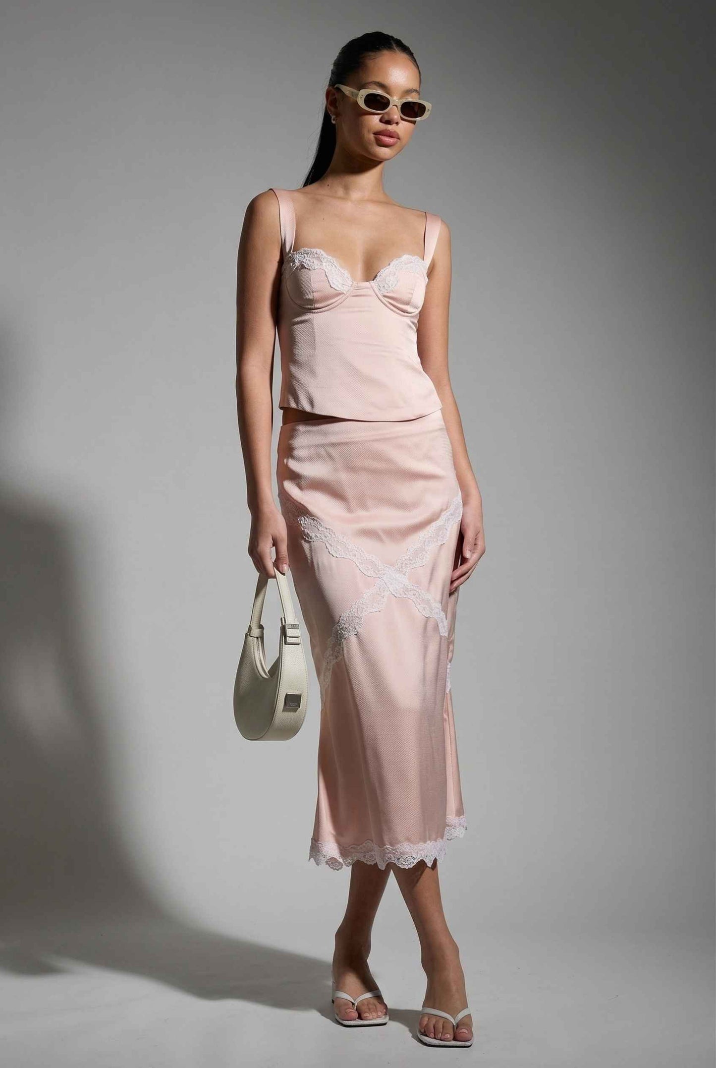 Woman wearing a Freya Skirt Blush Pink Satin Midi Skirt with White Lace Detail against a gray background