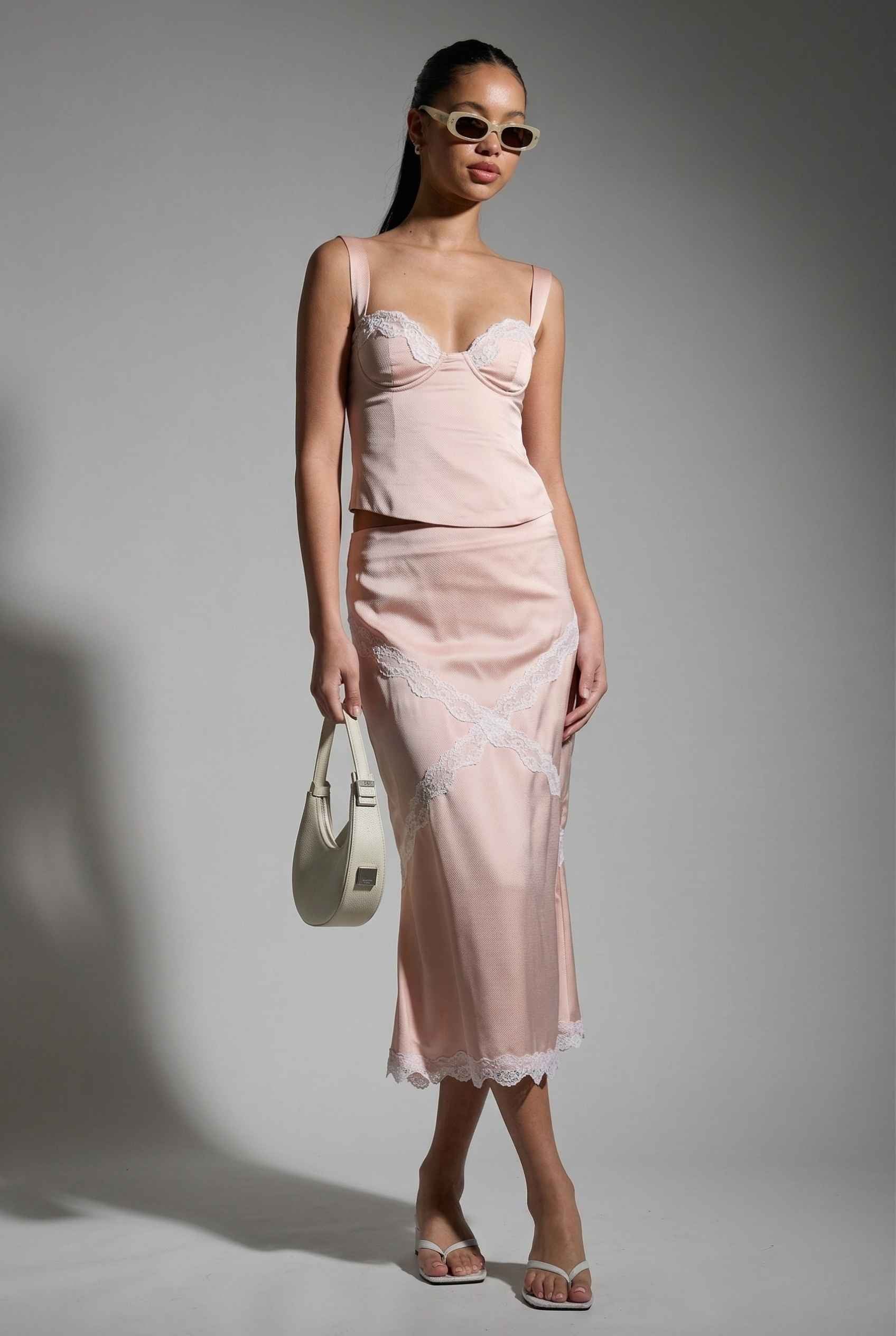 Woman wearing a Freya Skirt Blush Pink Satin Midi Skirt with White Lace Detail against a gray background