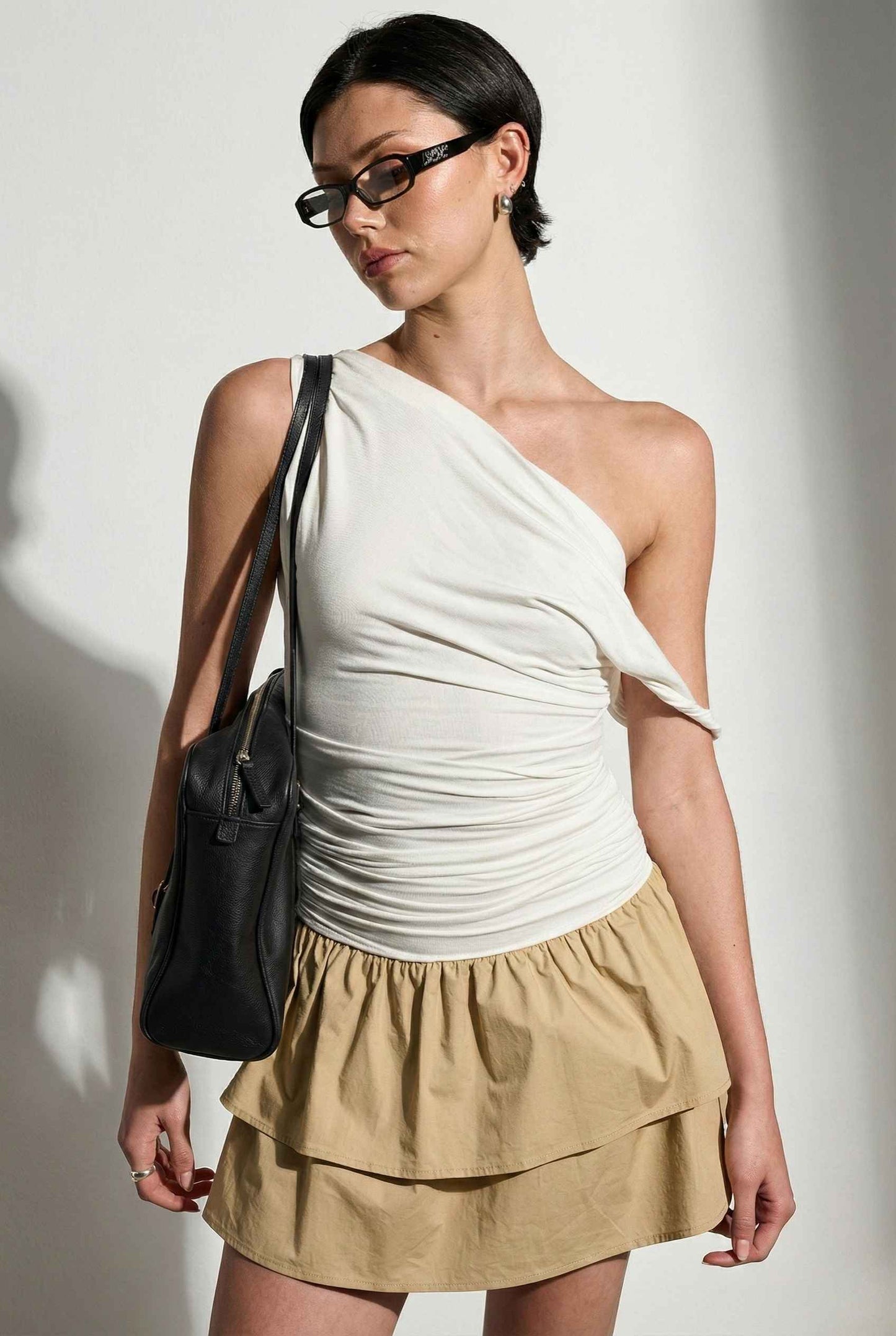 Woman wearing a Hadley Mini Ivory Ruched One-Shoulder Mixed Media Dress with a black bag against a plain background