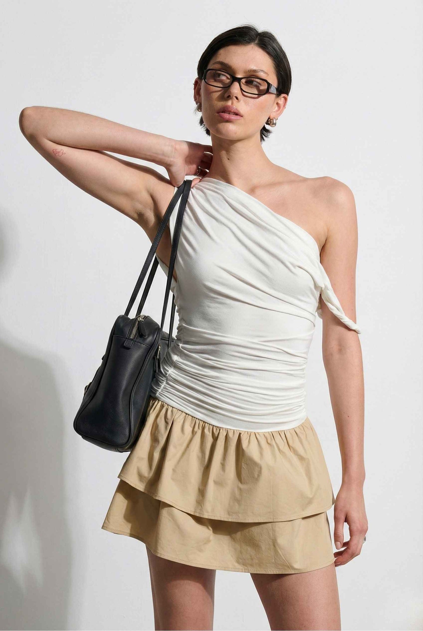 Woman wearing a Hadley Mini Ivory Ruched One-Shoulder Mixed Media Dress, holding a black handbag against a white background.