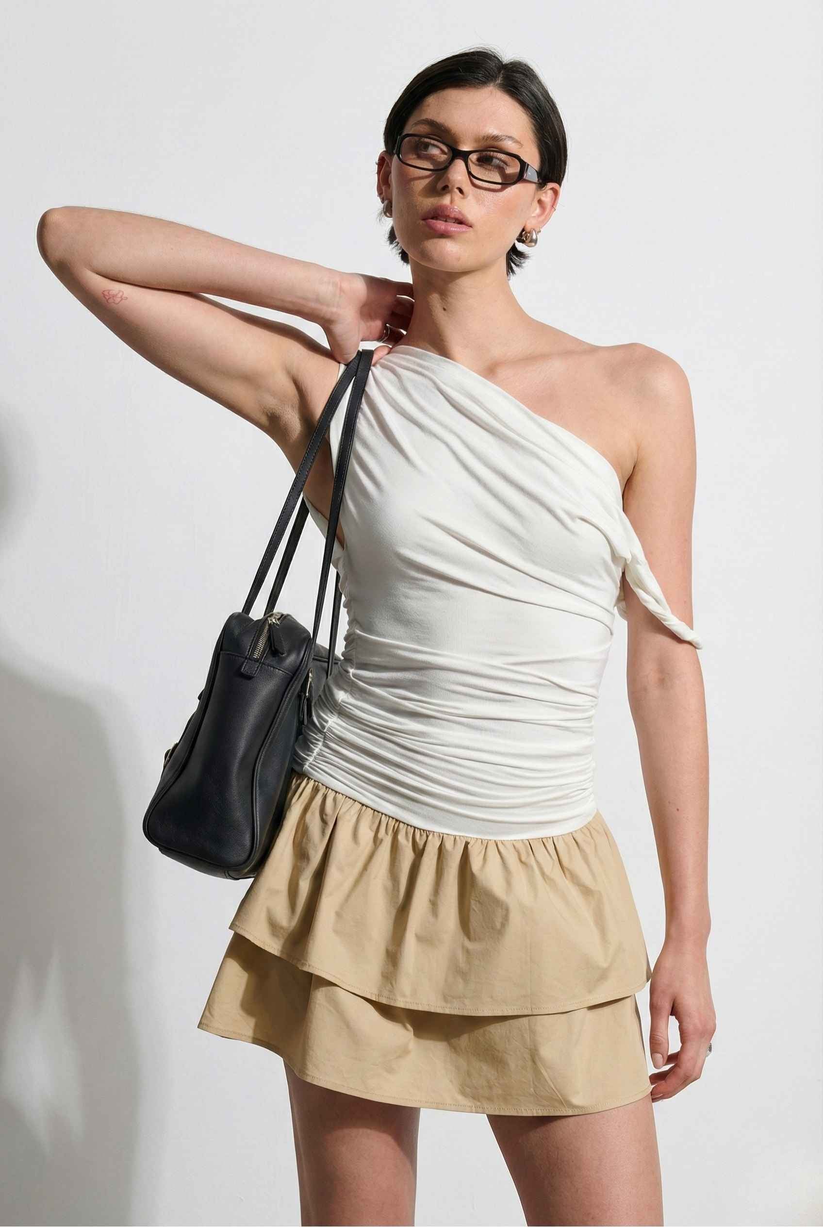 Woman wearing a Hadley Mini Ivory Ruched One-Shoulder Mixed Media Dress, holding a black handbag against a white background.
