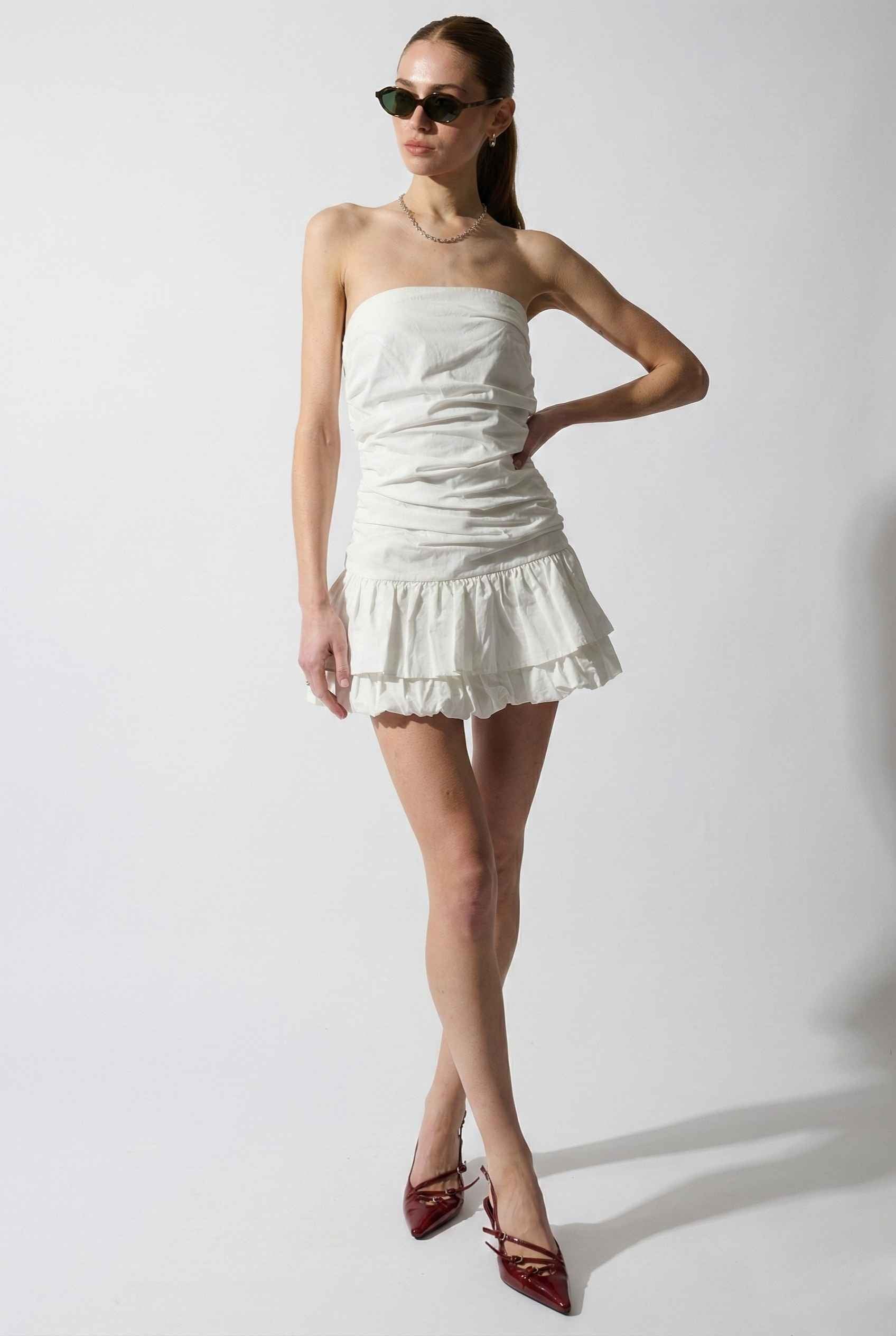 Woman wearing a Harper Mini White Ruched Strapless Cotton Dress with ruffled layers on a plain background