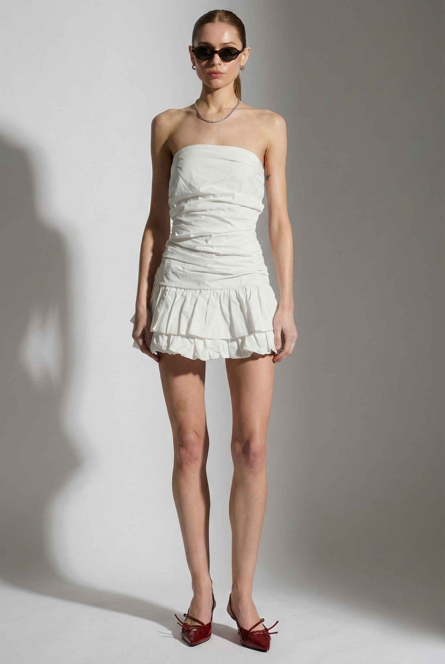 Woman wearing a Harper Mini White Ruched Strapless Cotton Dress with a ruffled skirt against a plain background