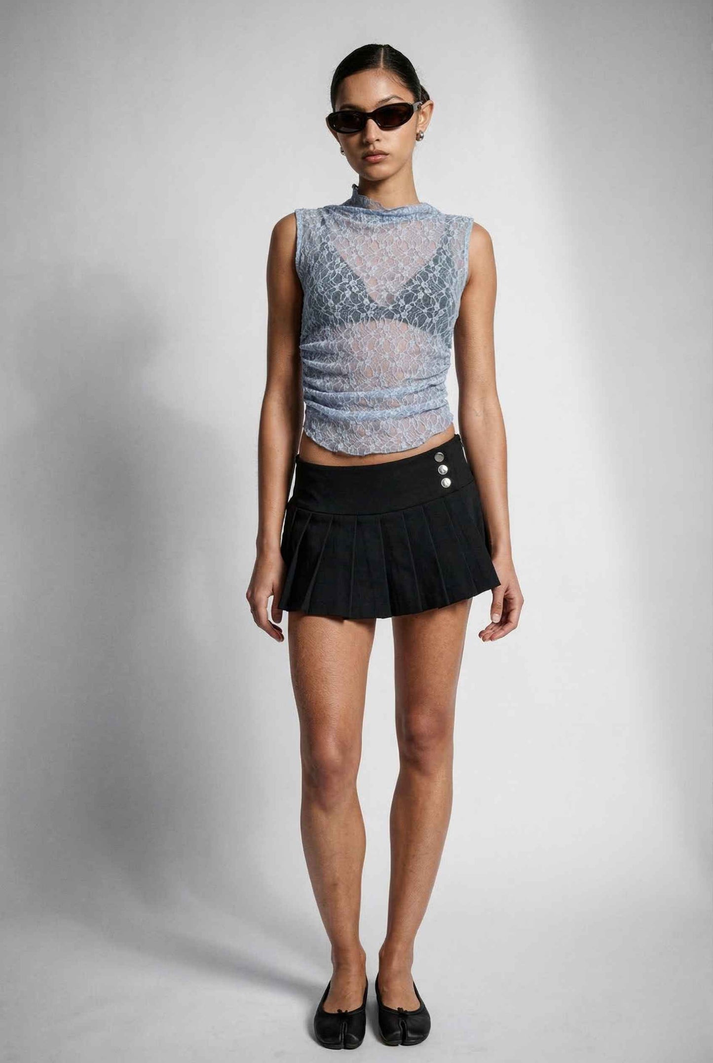 Woman wearing a Iris Top Dusty Blue Sheer Lace Sleeveless Mock Neck and black pleated skirt on a plain background