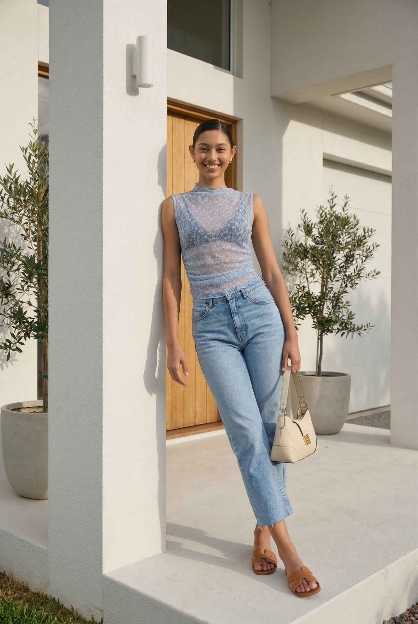 Woman in Iris Top Dusty Blue Sheer Lace Sleeveless Mock Neck and jeans standing in a modern outdoor setting.
