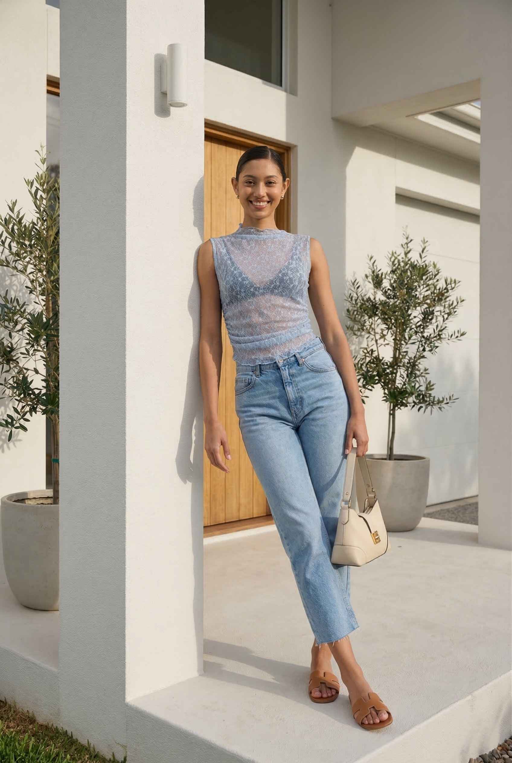 Woman in Iris Top Dusty Blue Sheer Lace Sleeveless Mock Neck and jeans standing in a modern outdoor setting.