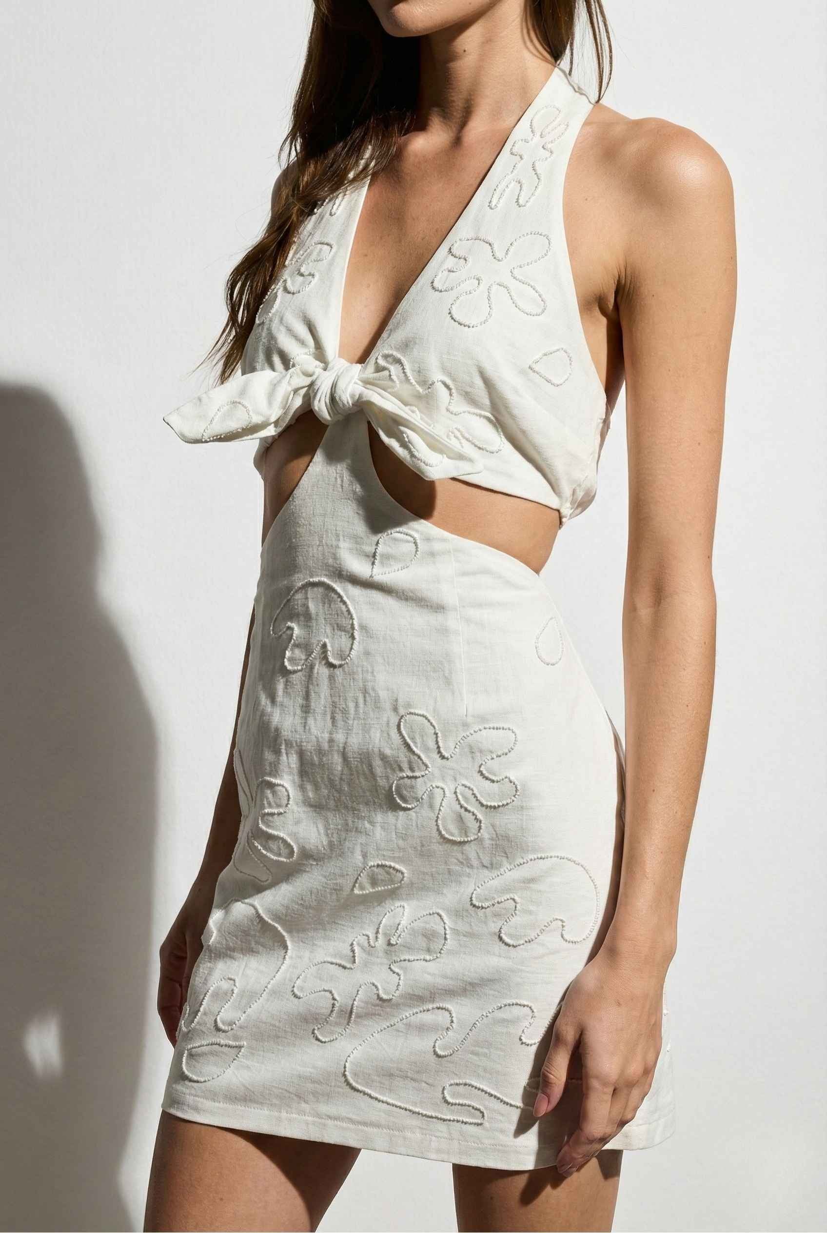 Woman wearing a Isla Mini White Linen Blend Halter Cut-Out Mini Dress with floral embroidery on a plain background
