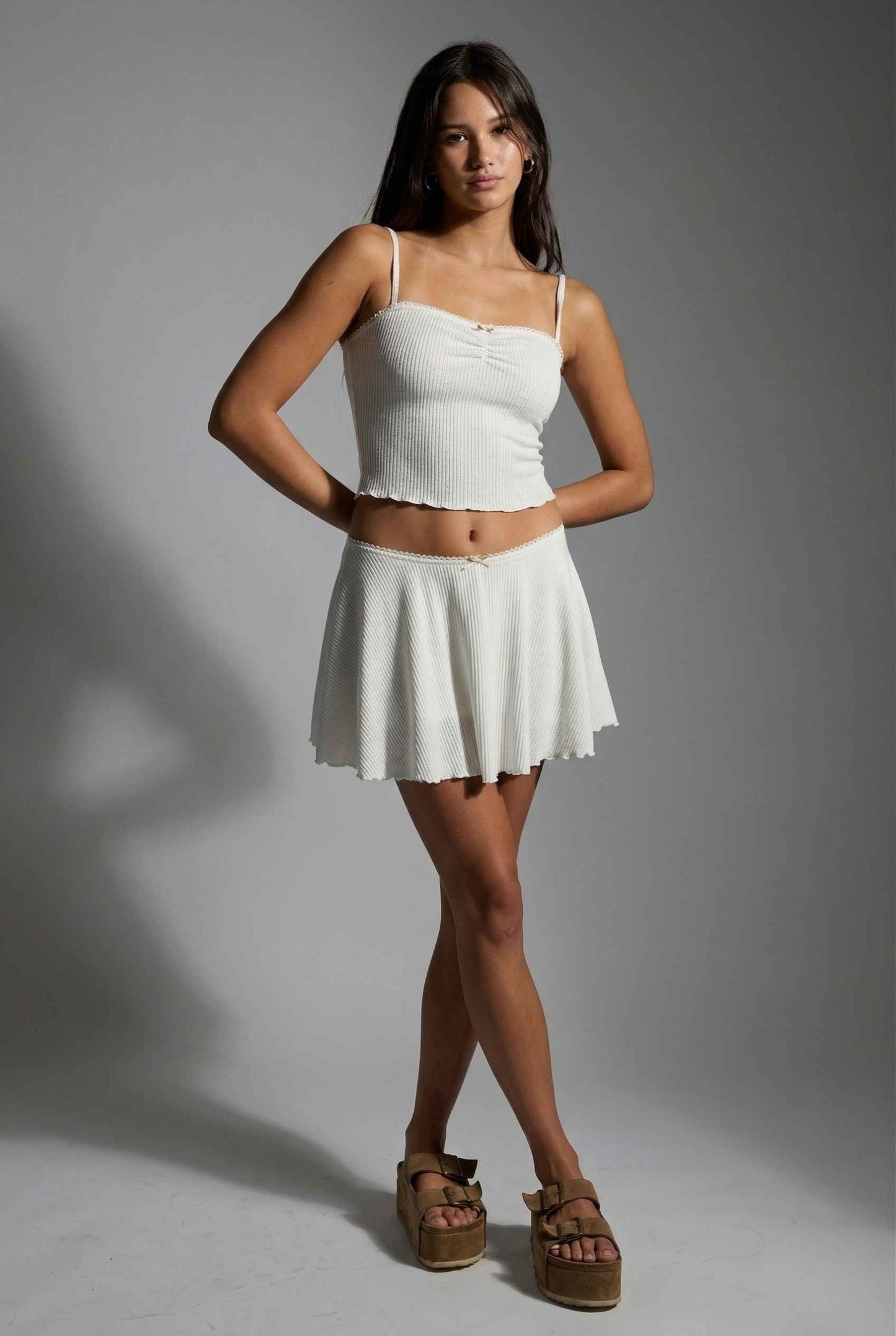 Woman wearing a white top and Ivy Skirt White Textured Knit Circle Mini Skirt with Bow with brown sandals on a gray background