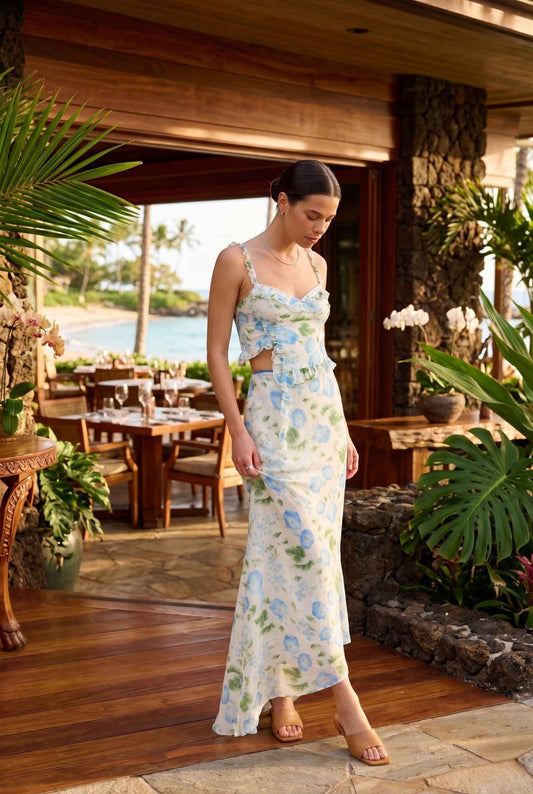 Woman in a Julia Asymmetrical Floral Bustier Top Ruffle Trim Top in Blue standing in a tropical outdoor setting with tables and chairs.