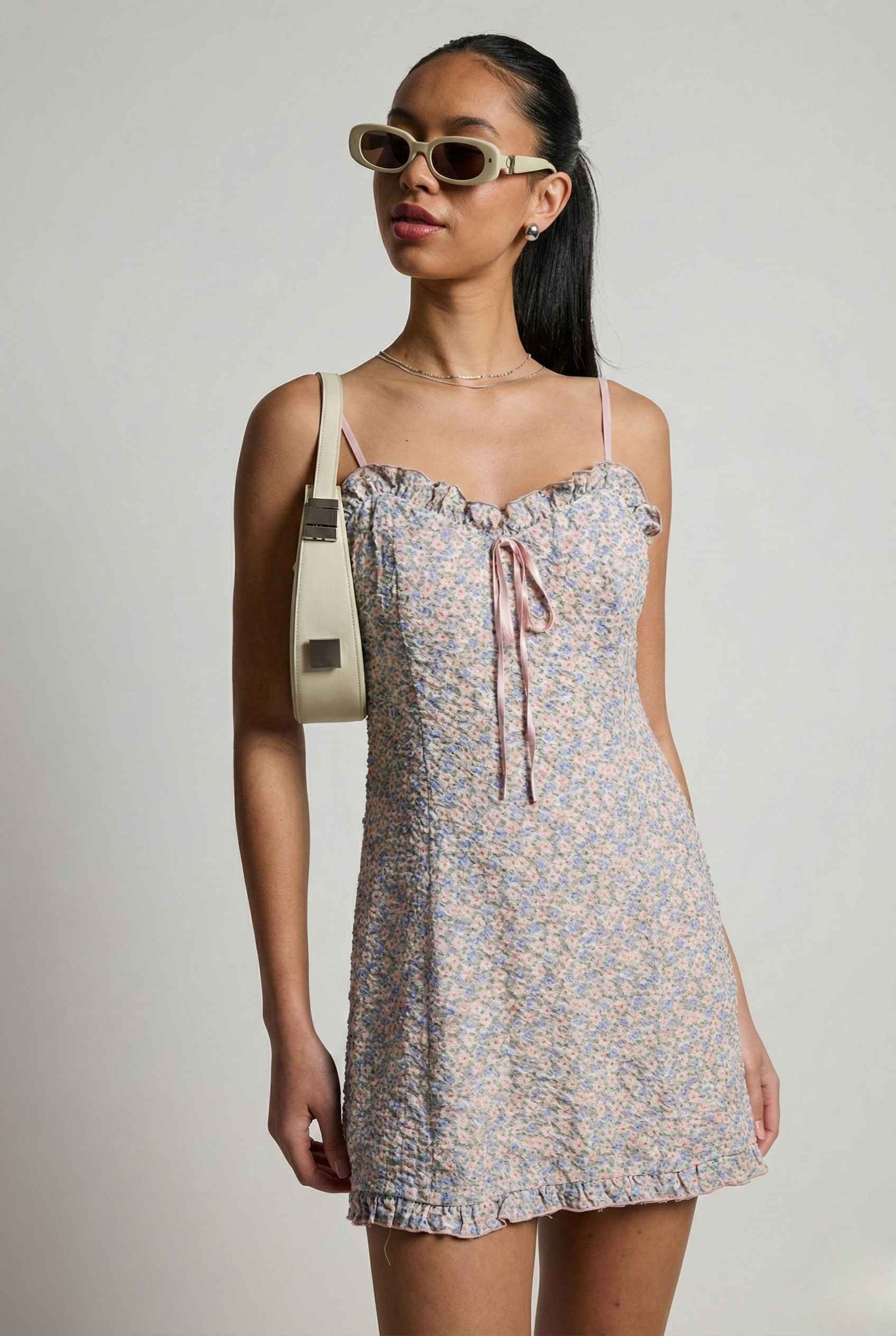 Woman wearing a Kaia Mini Pink Pastel Floral Ruffle Tie Dress with thin straps against a plain background