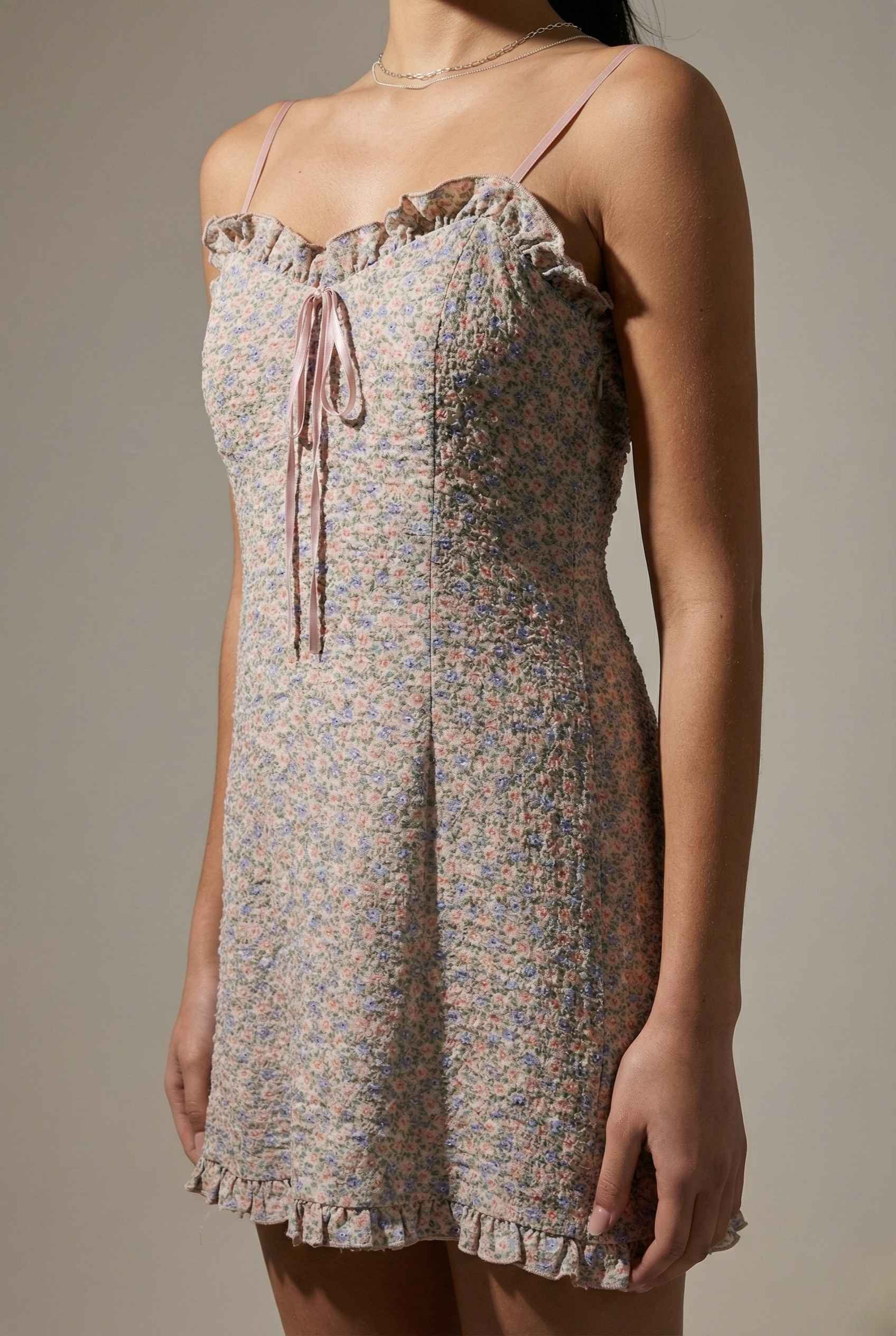 Woman wearing a Kaia Mini Pink Pastel Floral Ruffle Tie Dress with thin straps against a neutral background