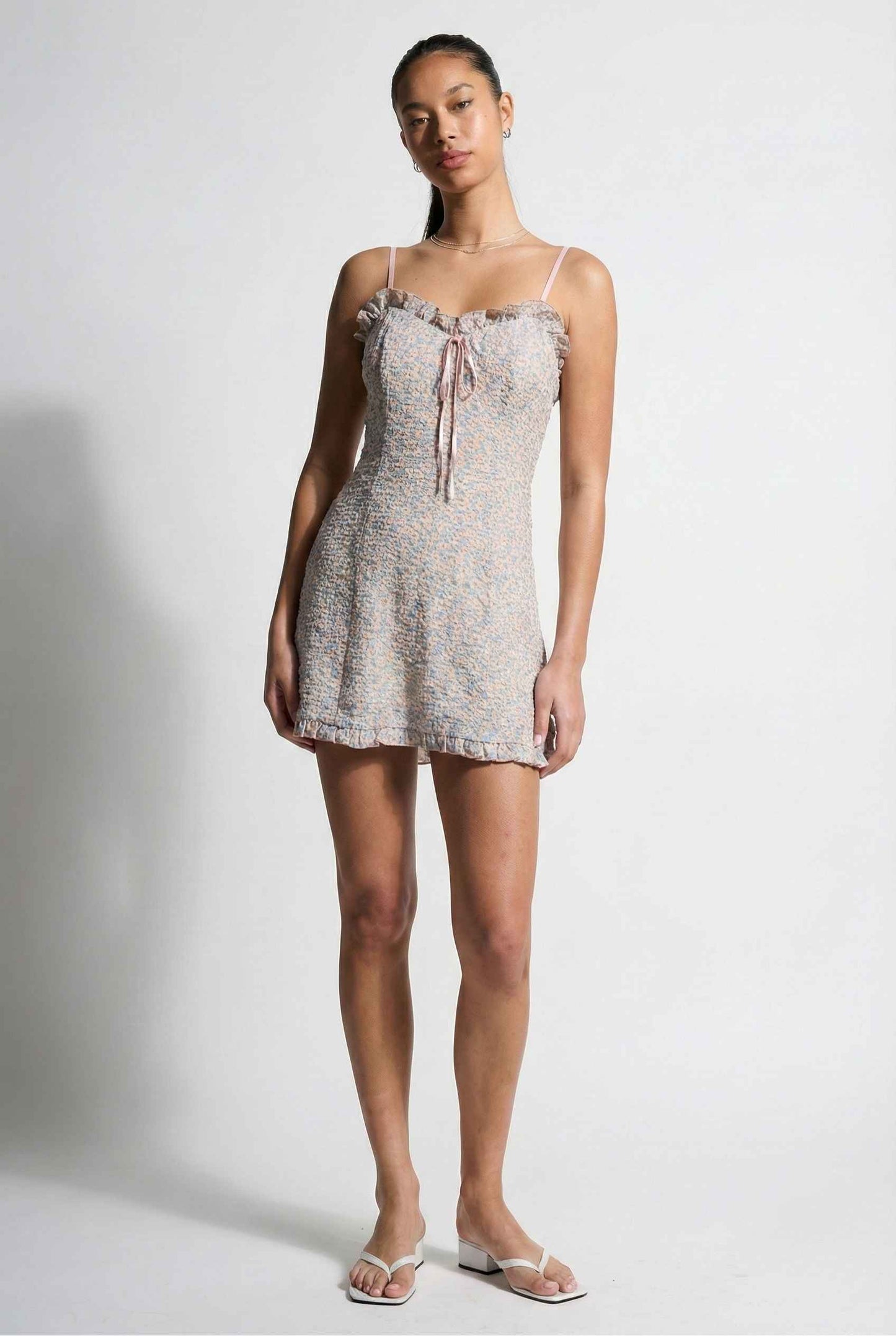 Woman wearing a Kaia Mini Pink Pastel Floral Ruffle Tie Dress with thin straps on a white background