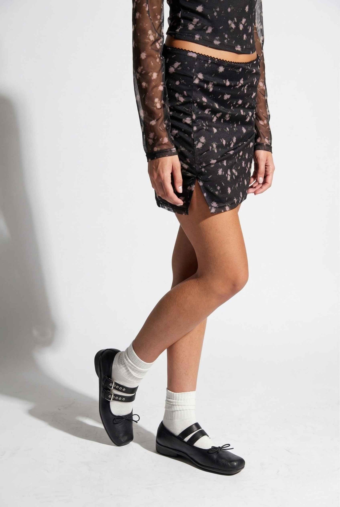 Woman wearing a Kaia Skirt Black Ditsy Floral Stretch Mesh Mini Skirt with sheer sleeves on a white background