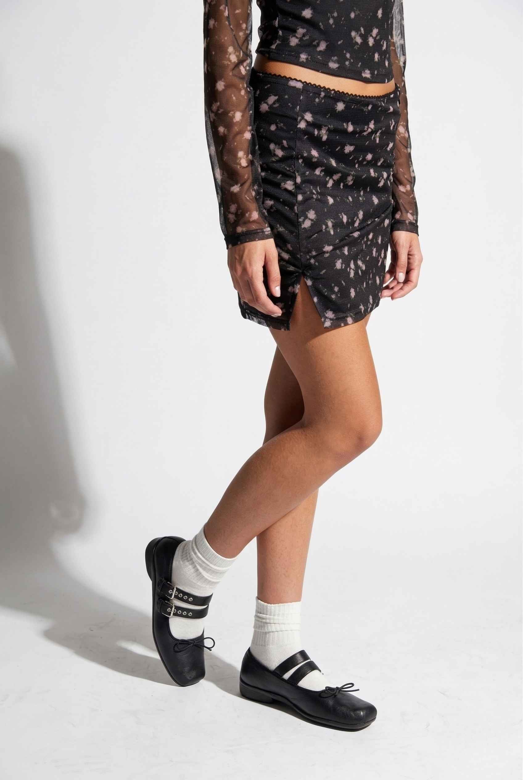 Woman wearing a Kaia Skirt Black Ditsy Floral Stretch Mesh Mini Skirt with sheer sleeves on a white background