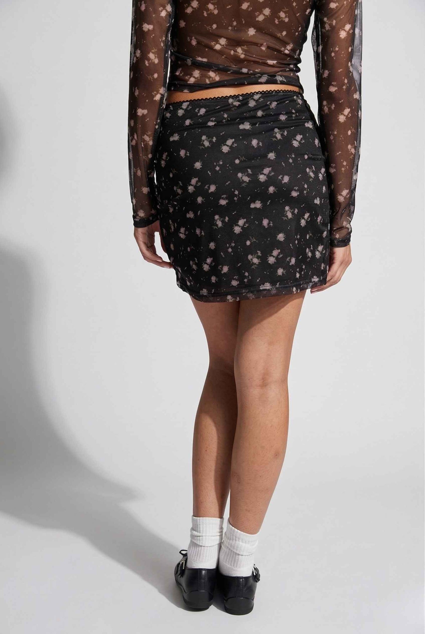 Woman wearing a Kaia Skirt Black Ditsy Floral Stretch Mesh Mini Skirt with sheer long-sleeve top on a white background