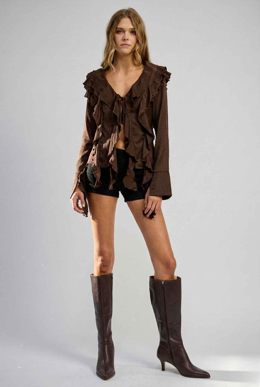 Woman wearing a Kaia Top Chocolate Brown Ruffle Tie-Front Long Sleeve Blouse and shorts with brown knee-high boots on a gray background