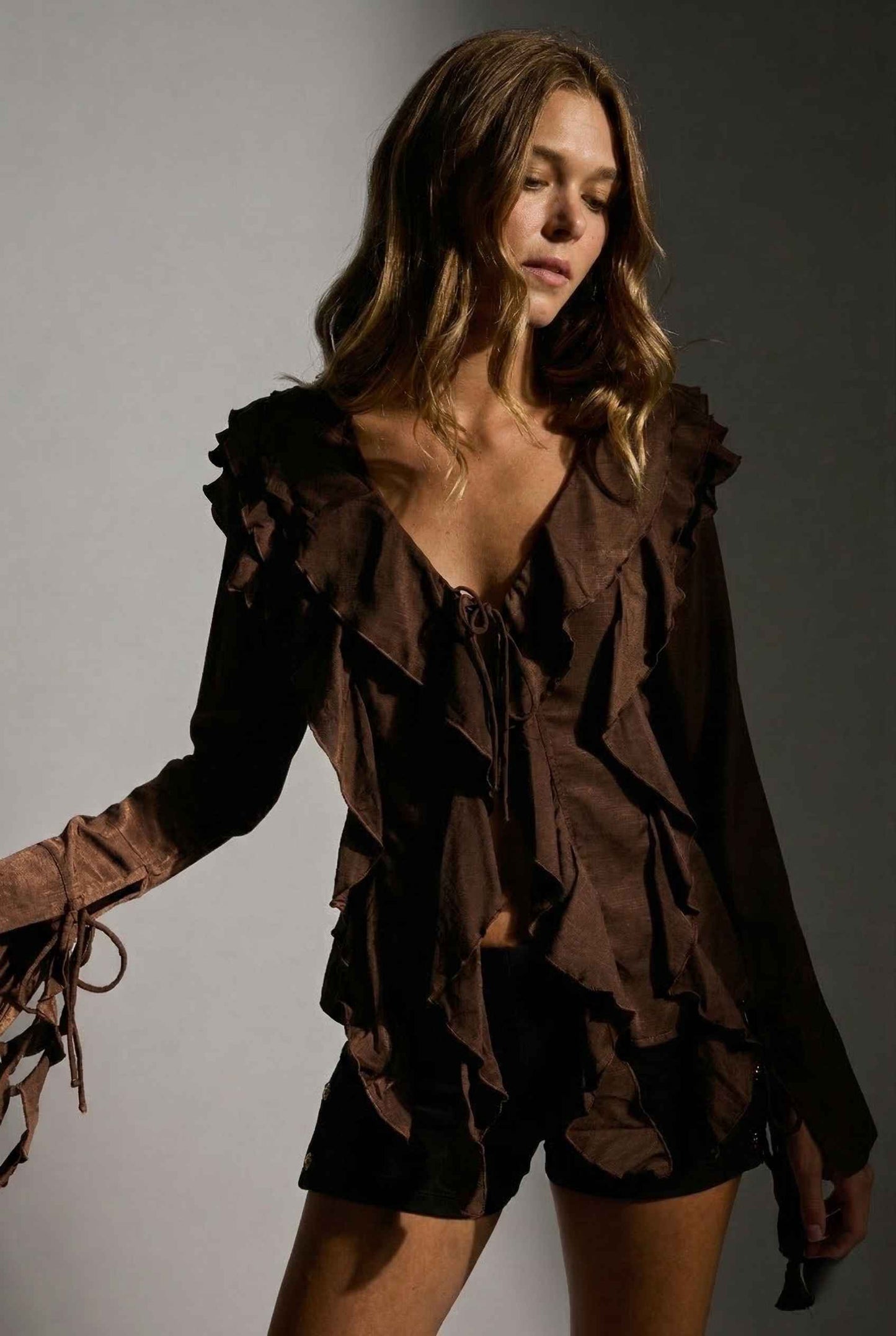 Woman wearing a Kaia Top Chocolate Brown Ruffle Tie-Front Long Sleeve Blouse and shorts against a gray background