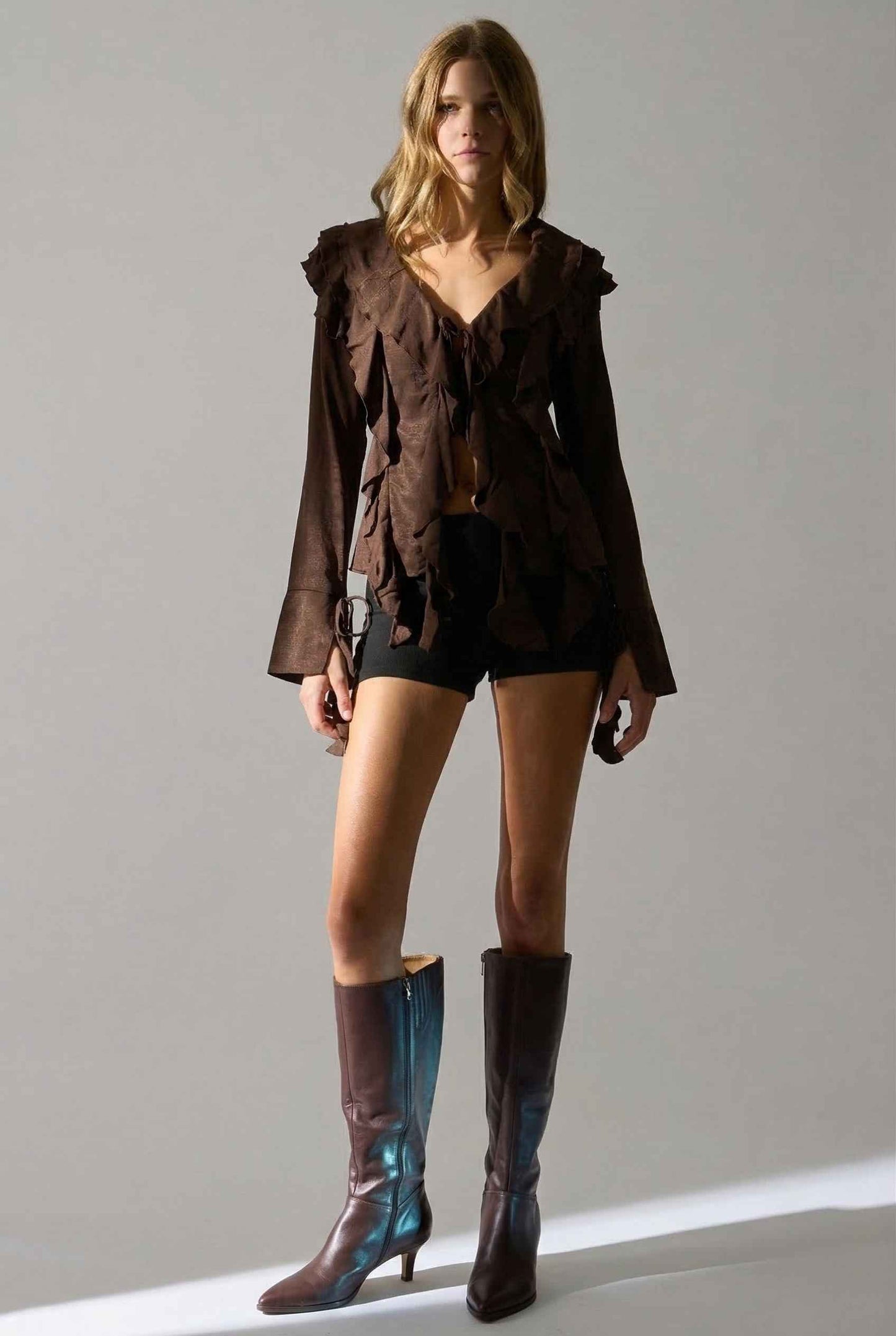 Woman wearing a Kaia Top Chocolate Brown Ruffle Tie-Front Long Sleeve Blouse with a jacket and boots on a plain background