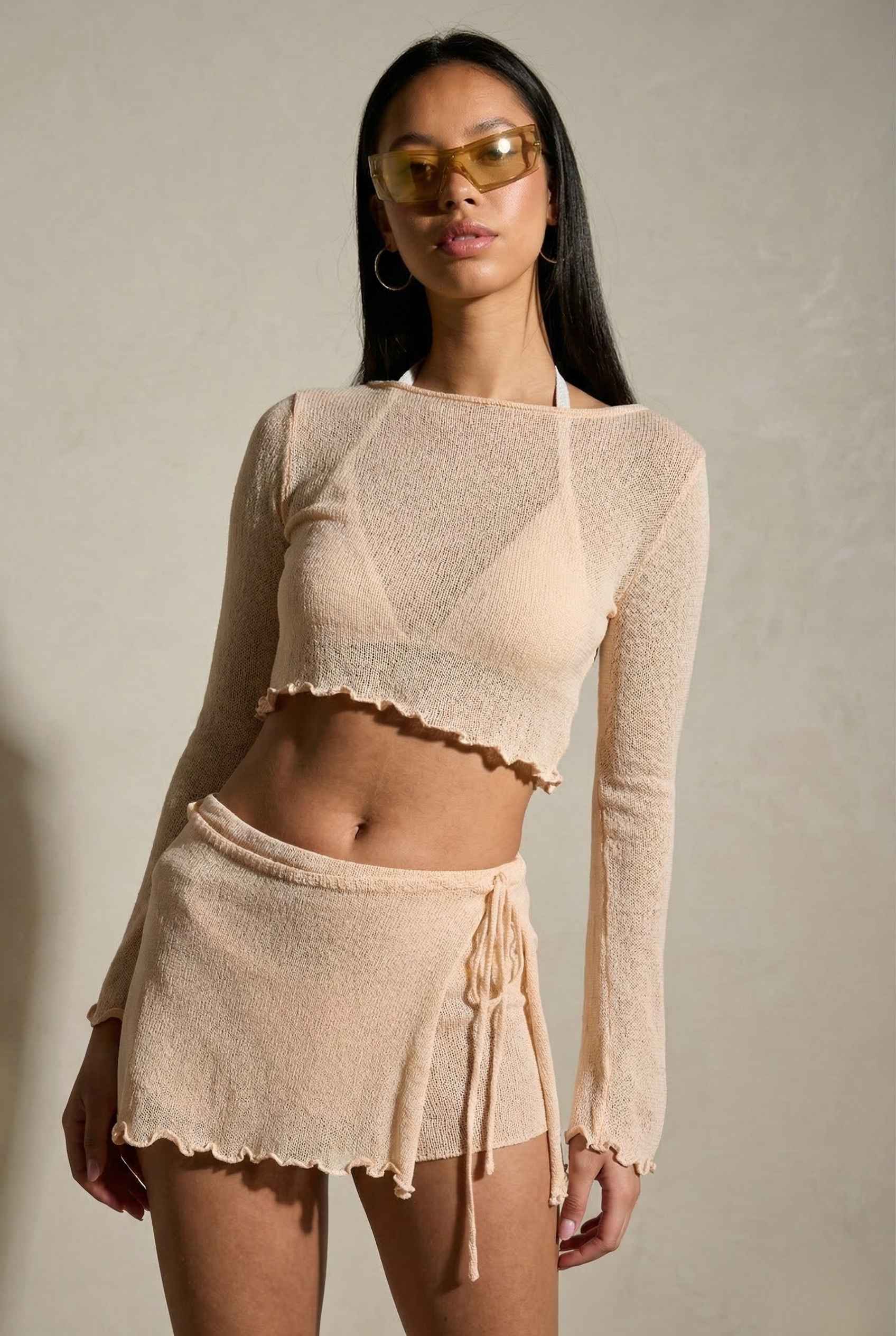 Woman wearing a Kaia Top Cream Sheer Knit Boat Neck Long Sleeve Crop with sunglasses against a plain background