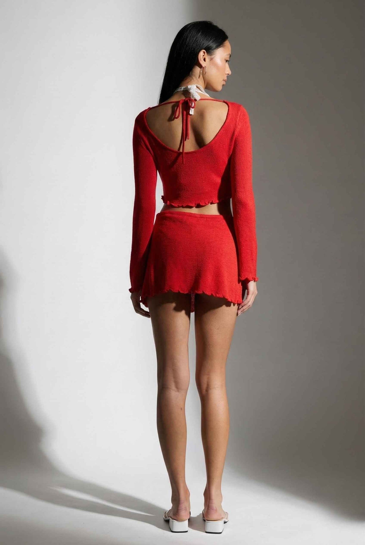 Woman wearing a Kaia Top Red Sheer Knit Boat Neck Long Sleeve Crop with a cut-out back design against a gray background