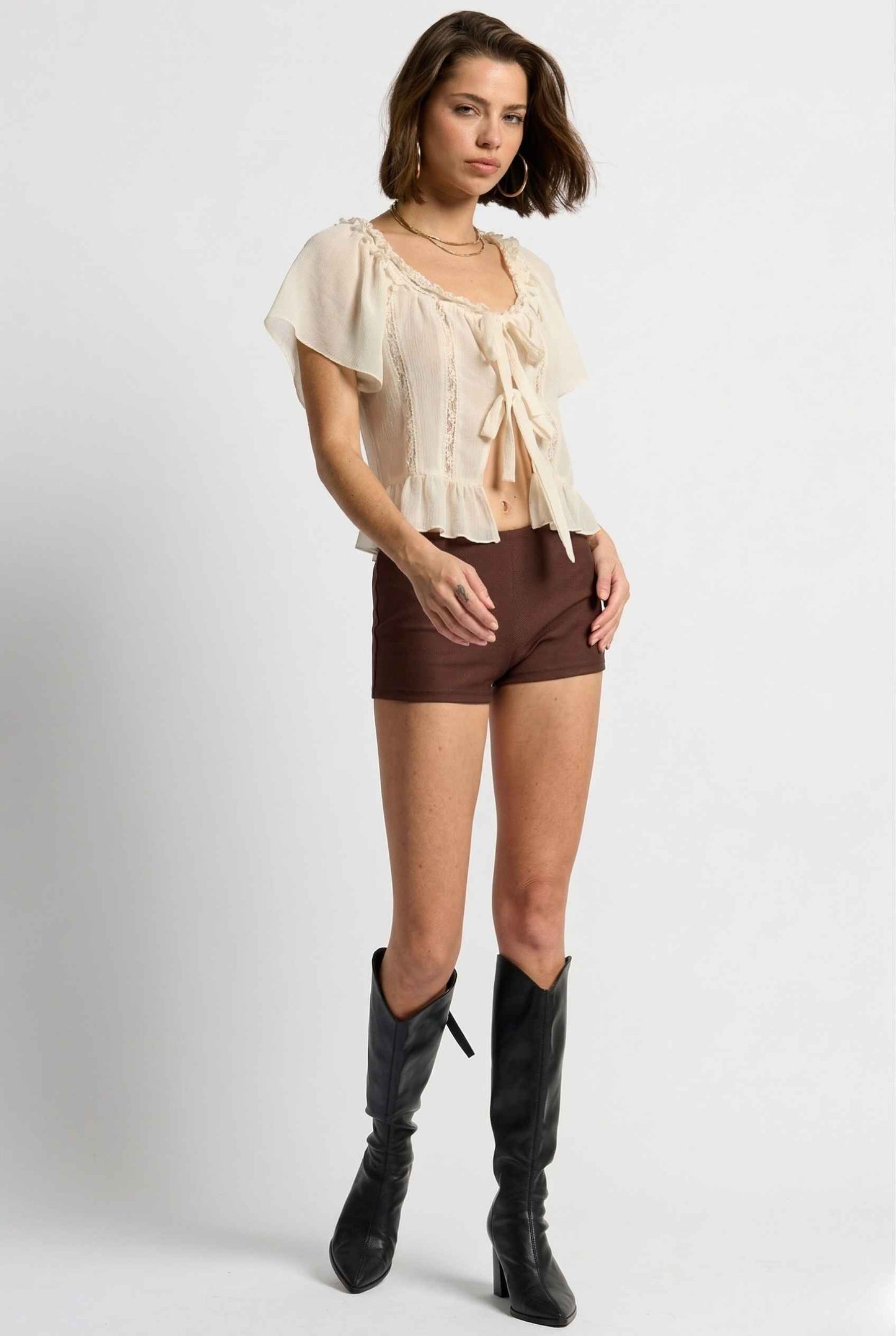 Woman wearing a beige blouse, Kyra Shorts Brown Rayon-Blend Knit Micro Biker Shorts, and knee-high boots on a white background